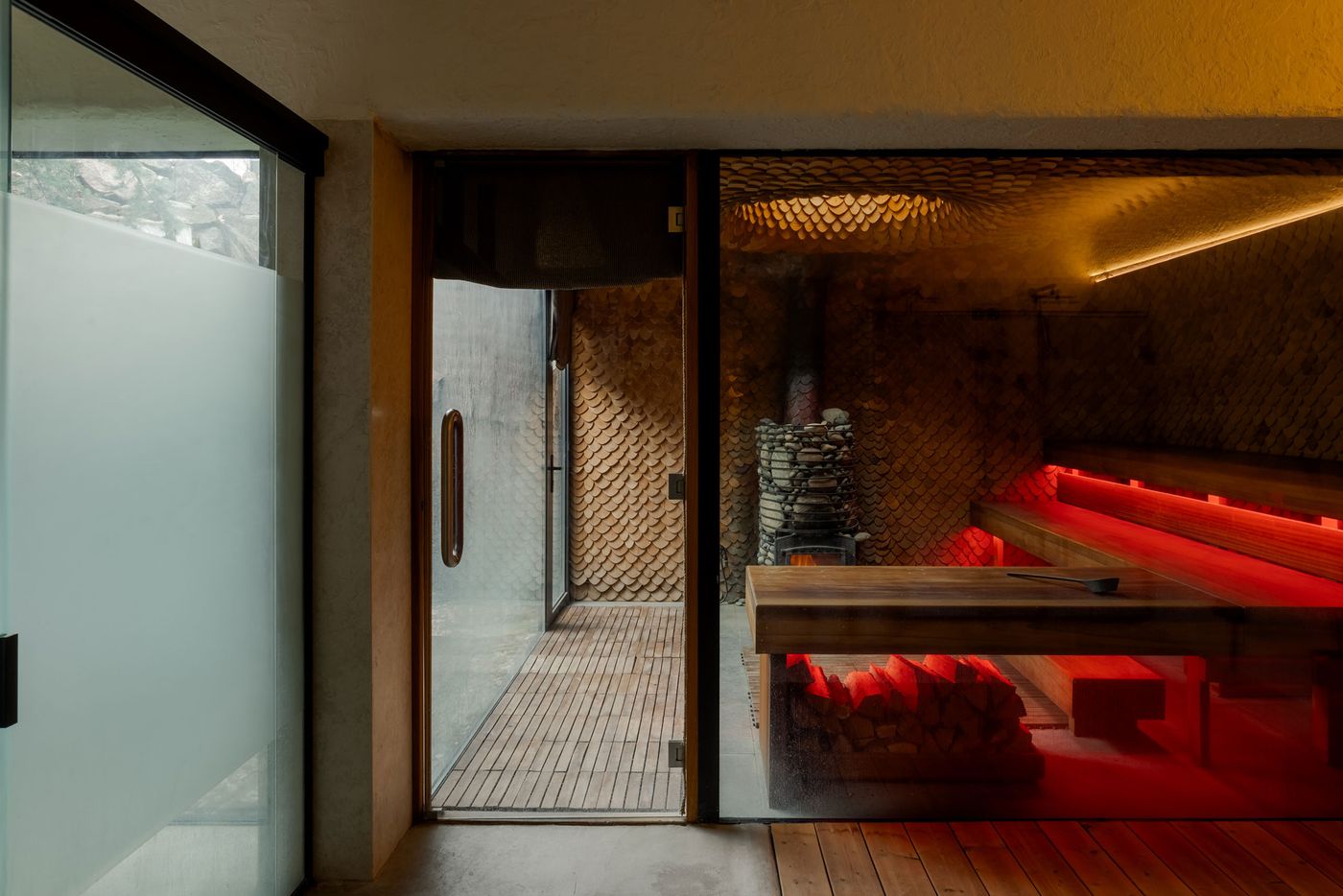 A modern relaxation space featuring wooden benches, warm red lighting, and a stone fireplace, with large glass doors revealing a natural view. A modern relaxation space featuring wooden benches, warm red lighting, and a stone fireplace, with large glass doors revealing a natural view.