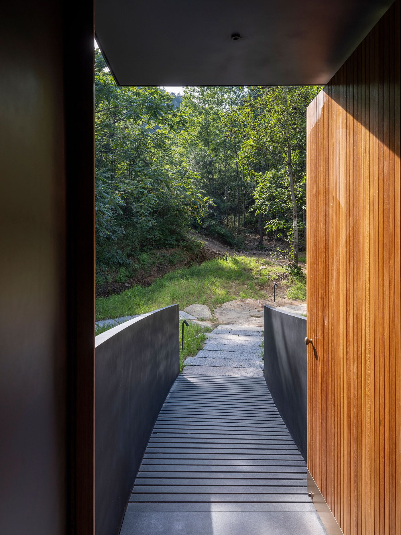 View from an open wooden door leading to a serene pathway through greenery, framed by trees and a gentle slope.