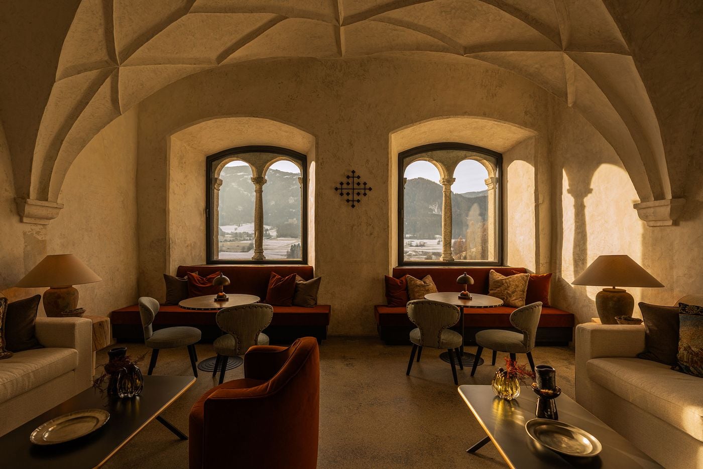 A Gothic net vault springs from carved corbels above the former Abbess's Hall at Castel Badia. Twin-arched stone windows frame the snow-covered valley beyond rust velvet banquettes and studded chairs, while cream sofas, terracotta lamps and a wrought-iron cross emblem create a symmetrical, sunlit composition. A Gothic net vault springs from carved corbels above the former Abbess's Hall at Castel Badia. Twin-arched stone windows frame the snow-covered valley beyond rust velvet banquettes and studded chairs, while cream sofas, terracotta lamps and a wrought-iron cross emblem create a symmetrical, sunlit composition.