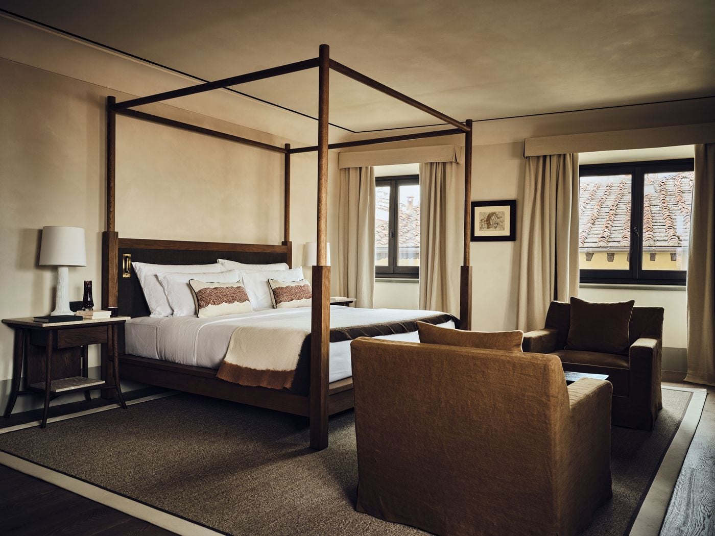 An oak four-poster bed anchors a bedroom at La Réserve Firenze, its windows dressed in linen with tailored pelmets and framing a patchwork of terracotta rooftops. A pair of rust linen armchairs, a stone-topped nightstand and a small framed architectural drawing keep the palette earthy and calm. An oak four-poster bed anchors a bedroom at La Réserve Firenze, its windows dressed in linen with tailored pelmets and framing a patchwork of terracotta rooftops. A pair of rust linen armchairs, a stone-topped nightstand and a small framed architectural drawing keep the palette earthy and calm.