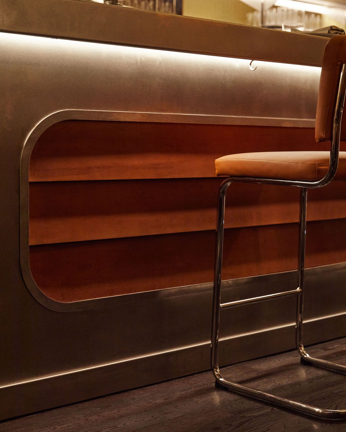 A detailed, low-angle shot of the bar counter. The sleek, brushed metal surface is softened by a recessed, curvilinear section of polished wood, adding a subtle warmth. A lone chrome-framed bar stool with a tan leather seat rests in front, highlighting the clean lines and meticulous craftsmanship of the space. A detailed, low-angle shot of the bar counter. The sleek, brushed metal surface is softened by a recessed, curvilinear section of polished wood, adding a subtle warmth. A lone chrome-framed bar stool with a tan leather seat rests in front, highlighting the clean lines and meticulous craftsmanship of the space.