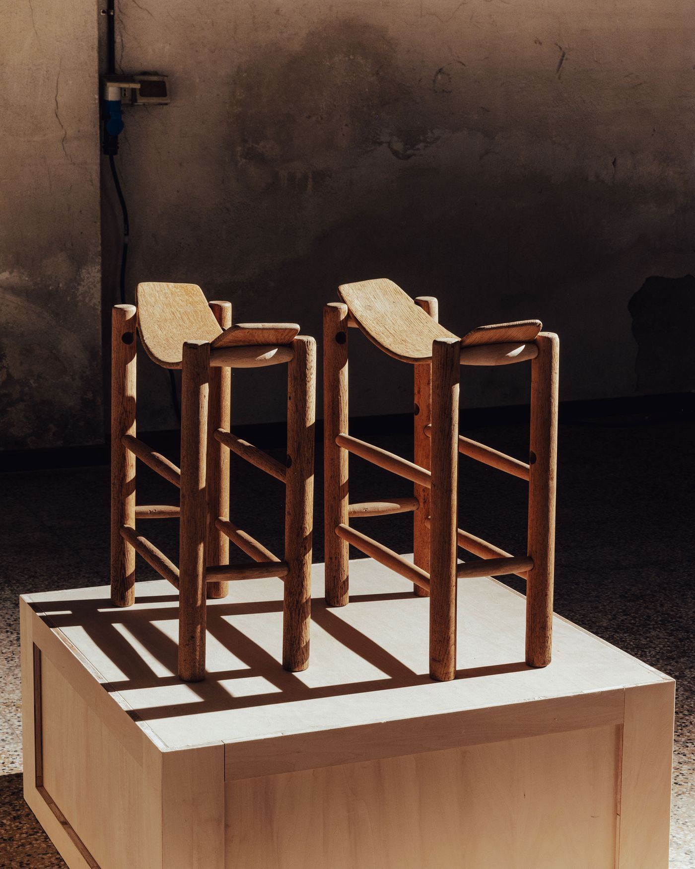 Two minimalist wooden stools with curved seats are displayed on a wooden pedestal, illuminated by soft sunlight against a textured wall. Two minimalist wooden stools with curved seats are displayed on a wooden pedestal, illuminated by soft sunlight against a textured wall.