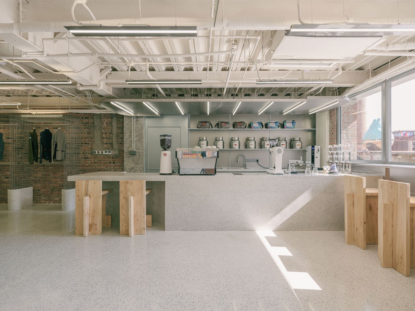 A pristine and functional cafe area features a long, light grey counter with integrated sinks and modern appliances, all neatly arranged on open shelving. Rustic wooden stools provide seating, contrasting with the polished terrazzo floor and exposed brick wall. Large windows flood the space with natural light, creating an inviting atmosphere. A pristine and functional cafe area features a long, light grey counter with integrated sinks and modern appliances, all neatly arranged on open shelving. Rustic wooden stools provide seating, contrasting with the polished terrazzo floor and exposed brick wall. Large windows flood the space with natural light, creating an inviting atmosphere.