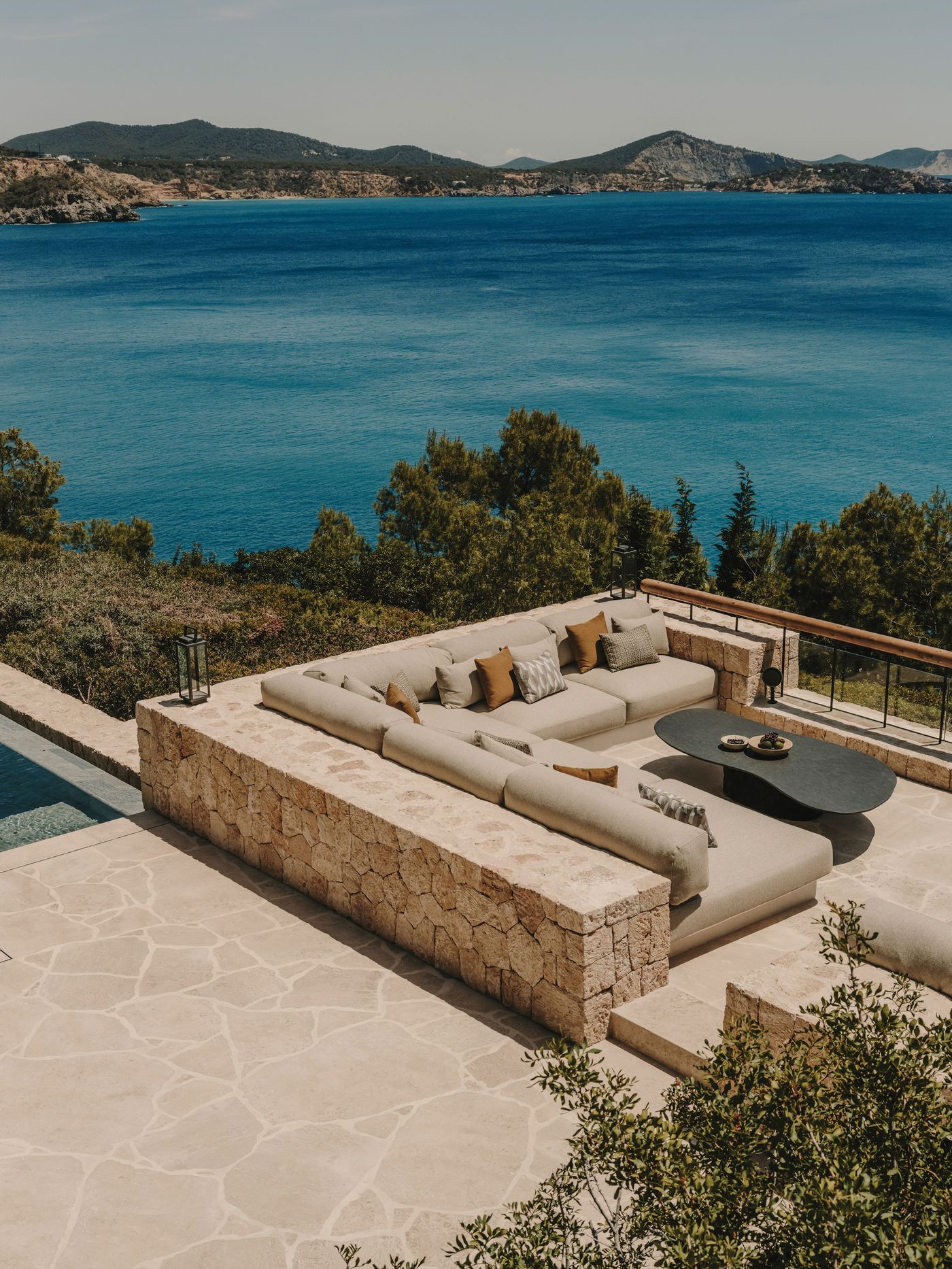 A modern outdoor lounge area with a stone wall, plush sofa, and table overlooks a serene blue sea and distant mountains under a clear sky.