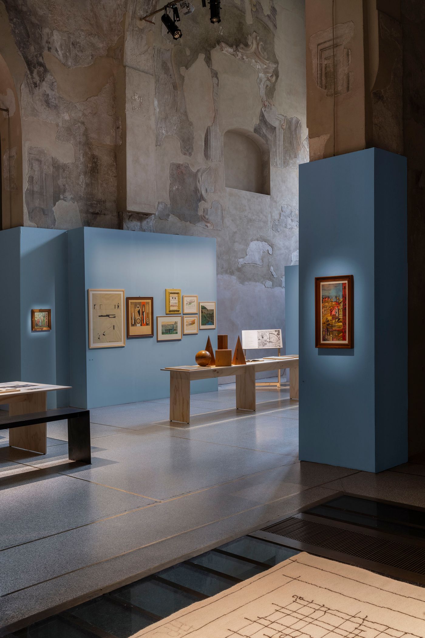 This exhibition space features light blue walls contrasting with the aged, textured surfaces of the historic hall. A large, detailed map-like artwork in warm golden tones is displayed prominently. Beside it, wooden tables present stacks of books and framed prints, alongside sculptural objects like cones and spheres. The composition emphasizes the interplay between contemporary display elements and the rich historical context of the venue.