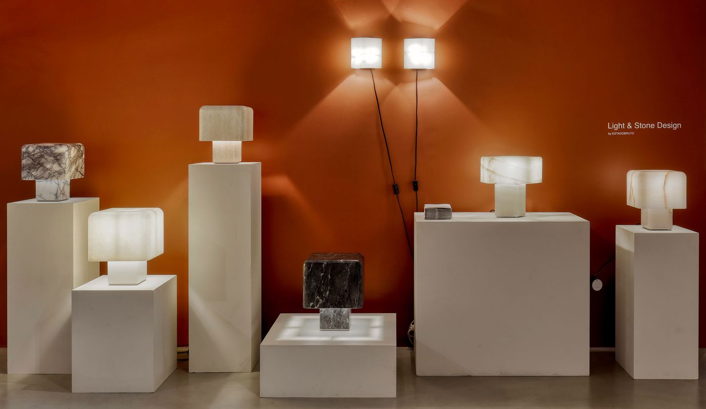A showroom display of "Light & Stone Design" by Estadeobruto Architects, featuring an array of minimalist stone lamps on various white pedestals against a vibrant orange wall. Translucent onyx and dark marble bases emit a warm, diffused glow, showcasing the interplay of natural materials and light. Wall-mounted rectangular sconces add to the sophisticated illumination of the display.