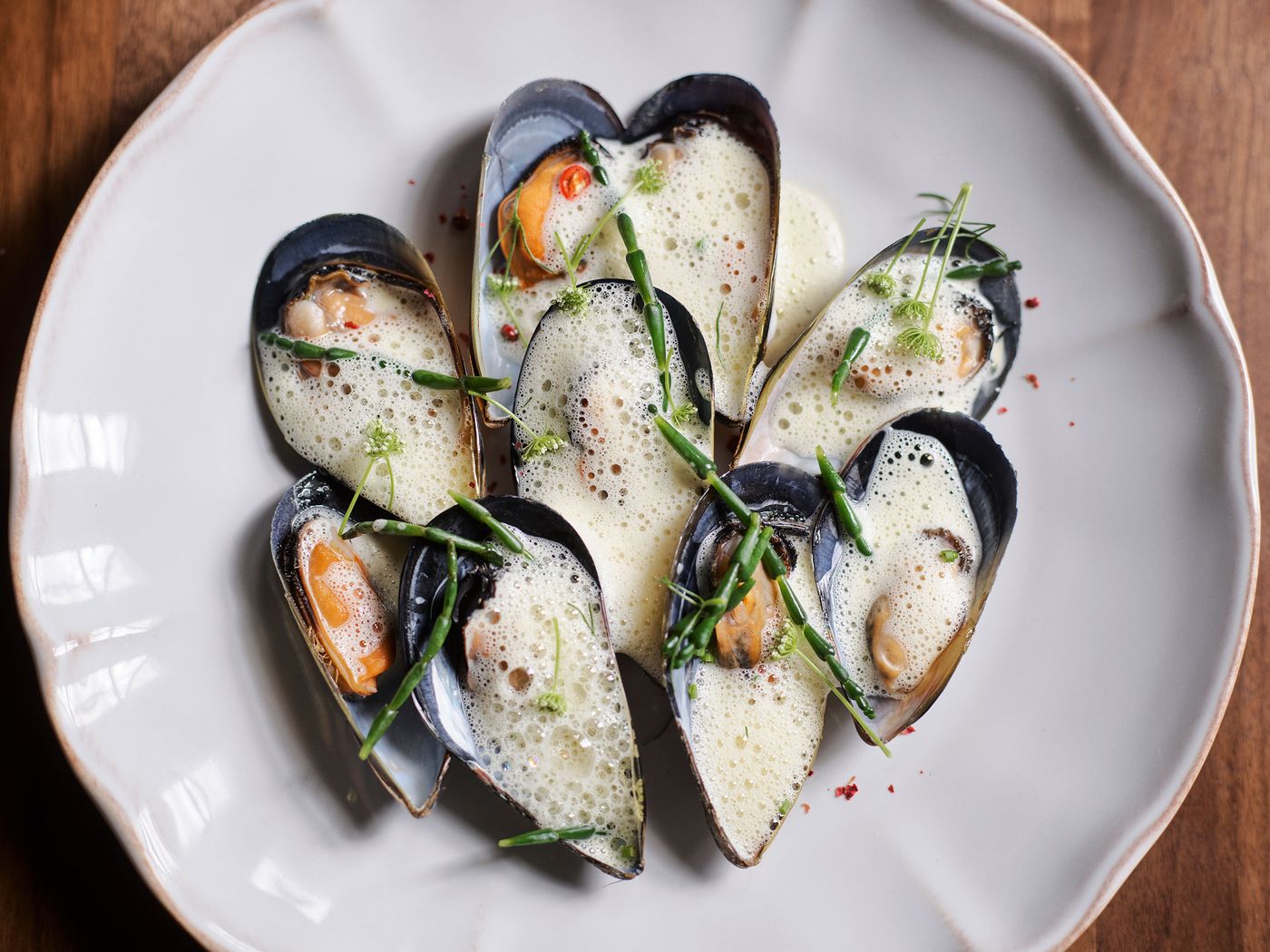 A white plate filled with mussels in black shells, topped with creamy foam and garnished with green chives and red pepper flakes. A white plate filled with mussels in black shells, topped with creamy foam and garnished with green chives and red pepper flakes.