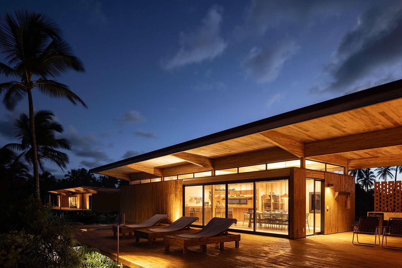 Under a vast twilight sky, a wood-clad structure glows with warm light from within, showcasing a seamless indoor-outdoor transition onto a sprawling wooden deck. Lounge chairs invite relaxation, while the palm trees beyond hint at a tranquil tropical setting.