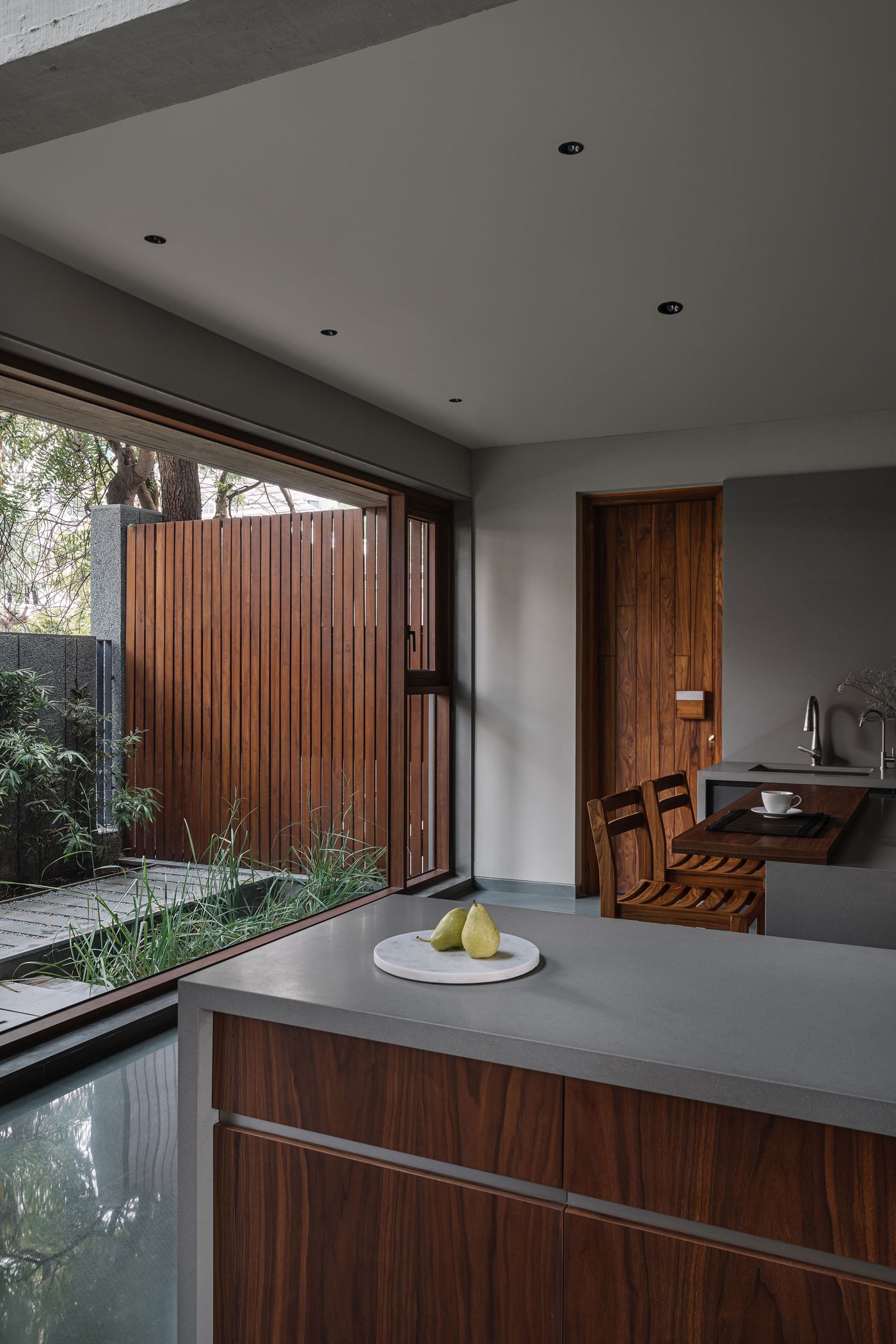 A minimalist kitchen and dining area opening onto an intimate outdoor courtyard through large sliding glass doors. The interior features sleek grey countertops and warm wooden cabinetry. The courtyard is defined by a slatted wooden fence and lush greenery, creating a private, biophilic oasis that blends seamlessly with the interior.