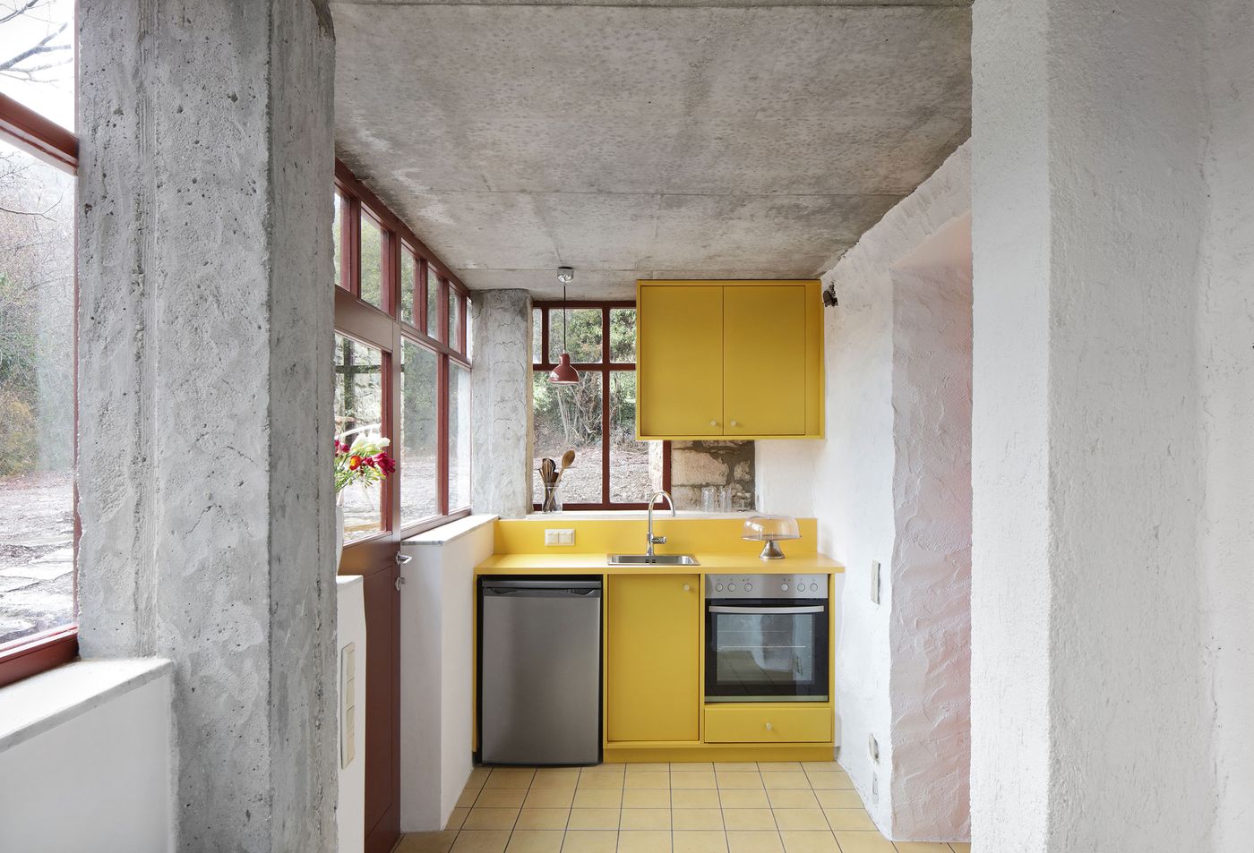Exposed concrete structural elements frame the efficient kitchen design, where yellow cabinetry provides cheerful contrast against the brutalist ceiling treatment. The minimal intervention demonstrates how contemporary elements can be inserted into historical buildings while preserving authentic architectural character and spatial relationships.