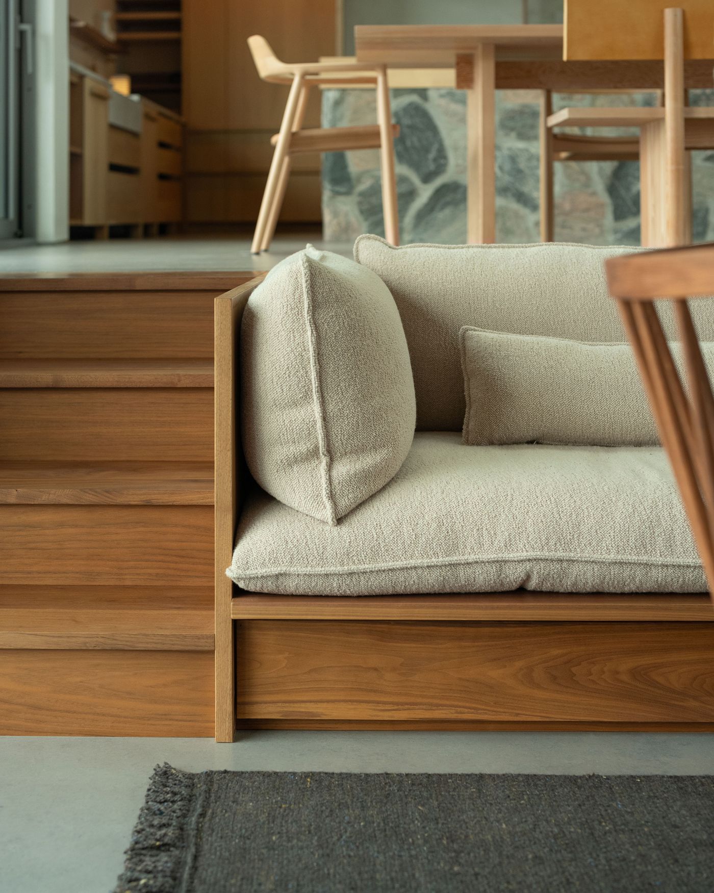 A cozy, modern wooden couch with soft beige cushions, positioned beside a textured rug and a dining area in a minimalist interior.