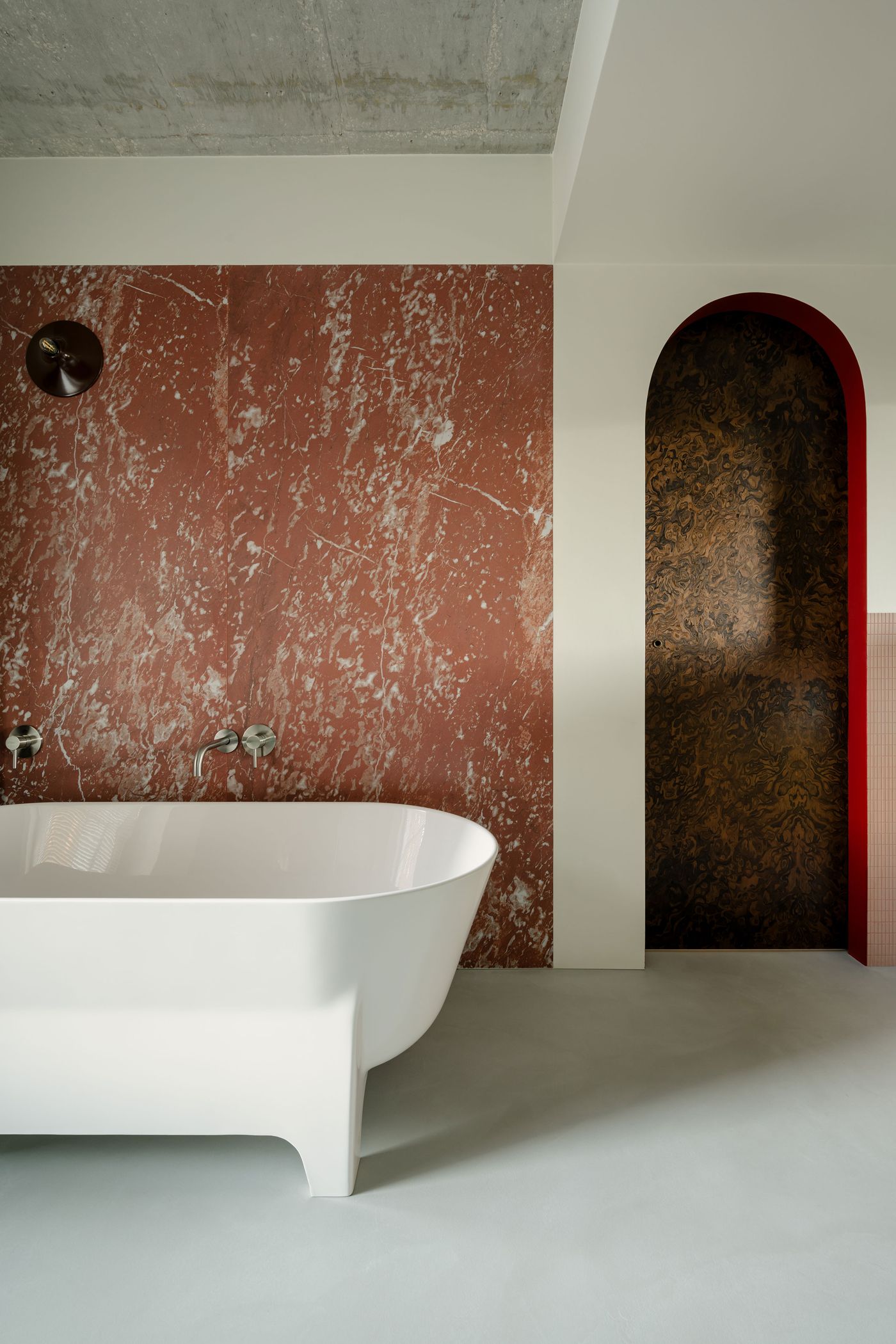 An architectural bathroom scene featuring a freestanding white tub, set against a rich red marble wall and framed by an arched walnut door. The tactile contrast of cool stone, warm wood, and soft curves adds depth to the minimalist layout. An architectural bathroom scene featuring a freestanding white tub, set against a rich red marble wall and framed by an arched walnut door. The tactile contrast of cool stone, warm wood, and soft curves adds depth to the minimalist layout.