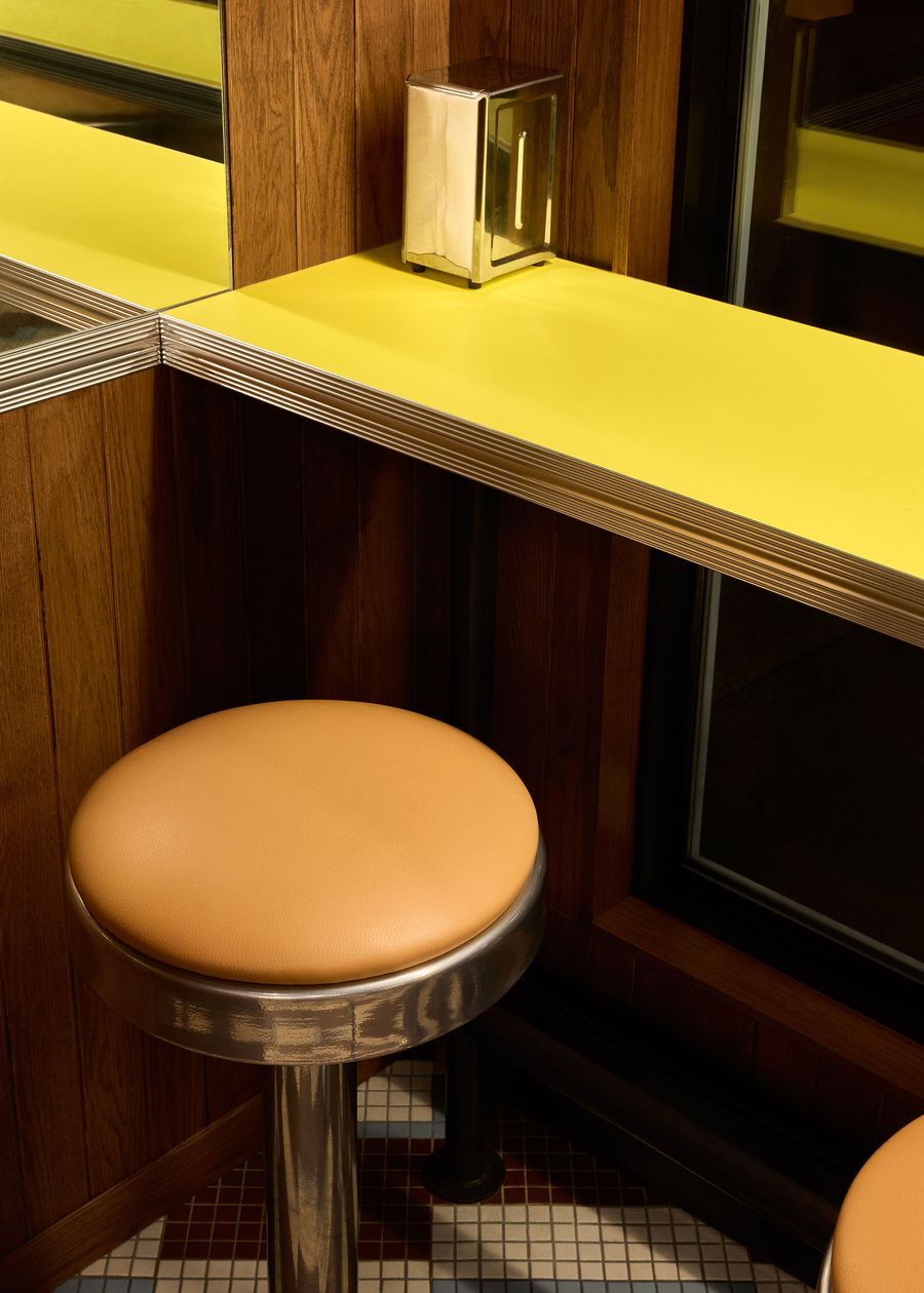 A close-up captures a detail of the Renzo Sandwich Shop interior, focusing on a sleek, tan leather stool with a chrome base positioned at a bright yellow counter. The counter, edged with polished metal, meets wood-paneled walls, and a vintage brass napkin dispenser sits on the surface. The geometric tile floor below adds to the establishment's retro charm.