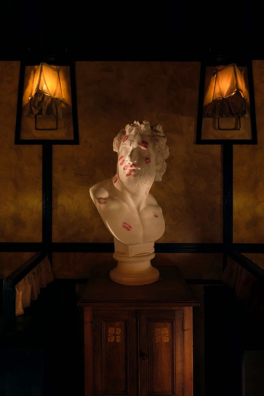 A playful yet sophisticated focal point at The Roses of Elagabalus: a classical white bust adorned with numerous red lipstick 'kisses,' placed atop a dark wooden cabinet. Two unique framed sconces with ruffled shades cast a warm glow on the textured, gold-hued walls, adding a touch of whimsical artistry to the vintage-inspired decor. A playful yet sophisticated focal point at The Roses of Elagabalus: a classical white bust adorned with numerous red lipstick 'kisses,' placed atop a dark wooden cabinet. Two unique framed sconces with ruffled shades cast a warm glow on the textured, gold-hued walls, adding a touch of whimsical artistry to the vintage-inspired decor.
