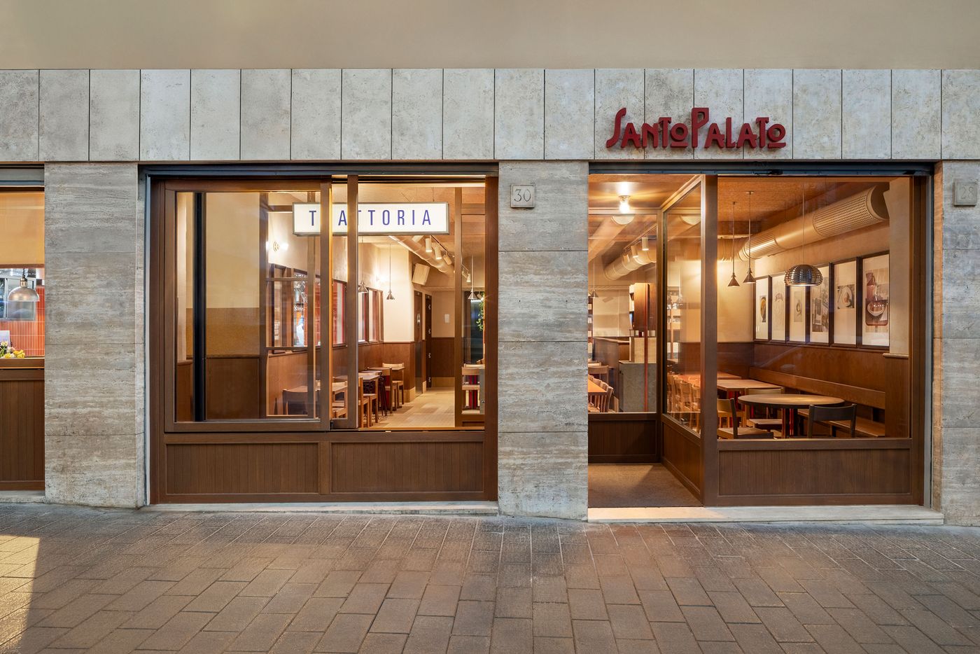 The inviting facade of Santo Palato, designed by Naessi Studio, is characterized by its modern travertine cladding and large wooden-framed glass windows. The illuminated "Santo Palato" sign above the entrance warmly beckons. Inside, glimpses of the vibrant orange communal table and the cozy dining areas promise an authentic Roman experience. The inviting facade of Santo Palato, designed by Naessi Studio, is characterized by its modern travertine cladding and large wooden-framed glass windows. The illuminated "Santo Palato" sign above the entrance warmly beckons. Inside, glimpses of the vibrant orange communal table and the cozy dining areas promise an authentic Roman experience.