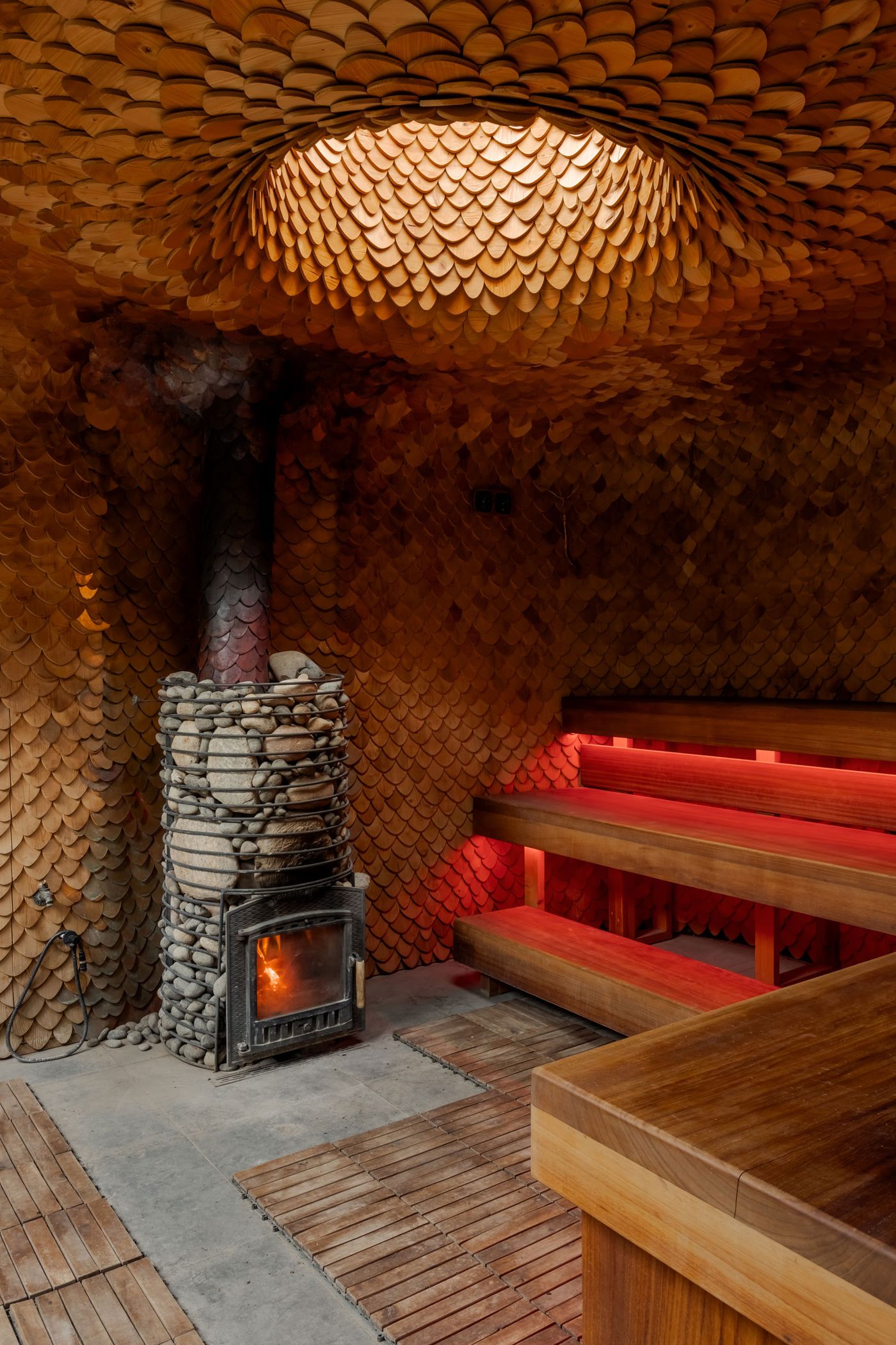 Interior of a cozy, earthy sauna featuring a stone fireplace, wooden benches, and intricate wooden scales on the ceiling and walls.