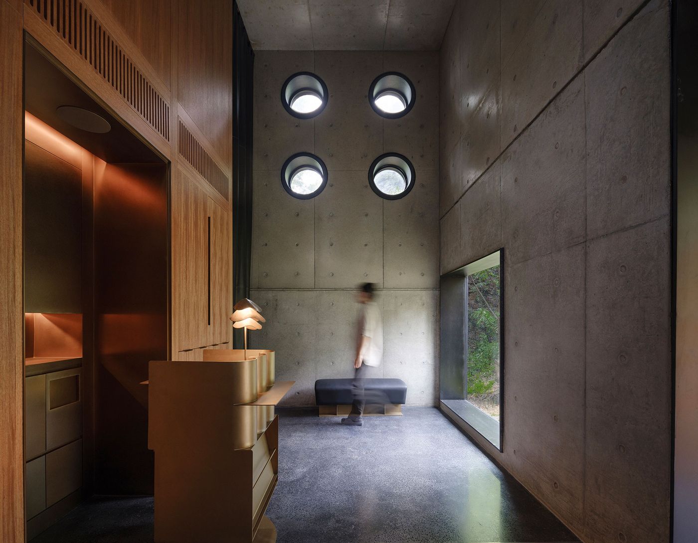A minimalist interior with concrete walls, circular windows, and a person walking past a sleek maple wood counter and large side window. A minimalist interior with concrete walls, circular windows, and a person walking past a sleek maple wood counter and large side window.