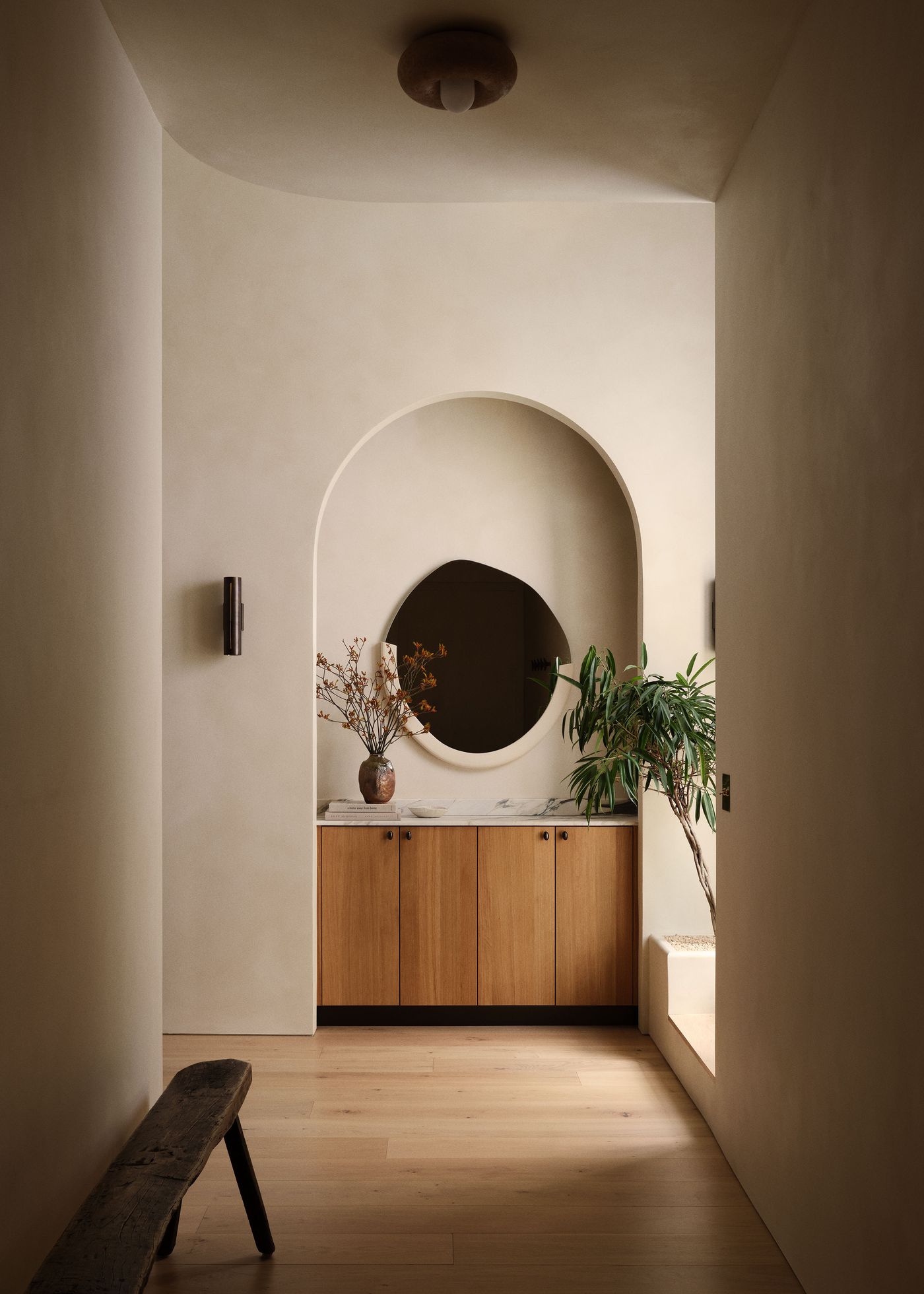 Minimalist hallway featuring a wooden cabinet, round mirror, potted plants, and natural light, with soft beige walls and wooden flooring.