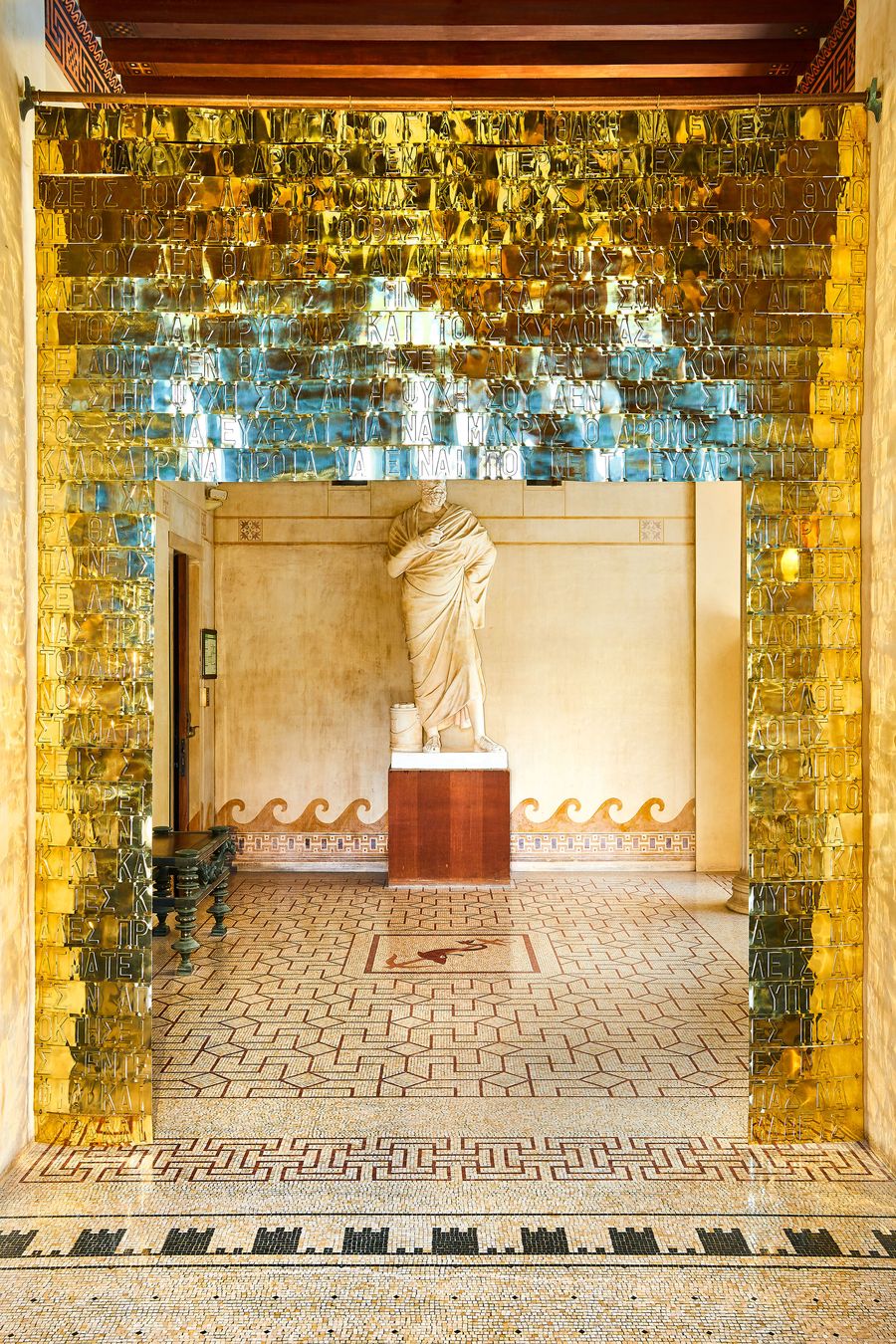 A head-on view of Gabriel Leger's sculptural installation at Villa Kerylos. The cascade of golden metallic sheets, etched with Greek script, creates a translucent doorway. Beyond, a classical statue stands in a sun-drenched corridor with intricate wall friezes and a subtle floor mosaic. A head-on view of Gabriel Leger's sculptural installation at Villa Kerylos. The cascade of golden metallic sheets, etched with Greek script, creates a translucent doorway. Beyond, a classical statue stands in a sun-drenched corridor with intricate wall friezes and a subtle floor mosaic.