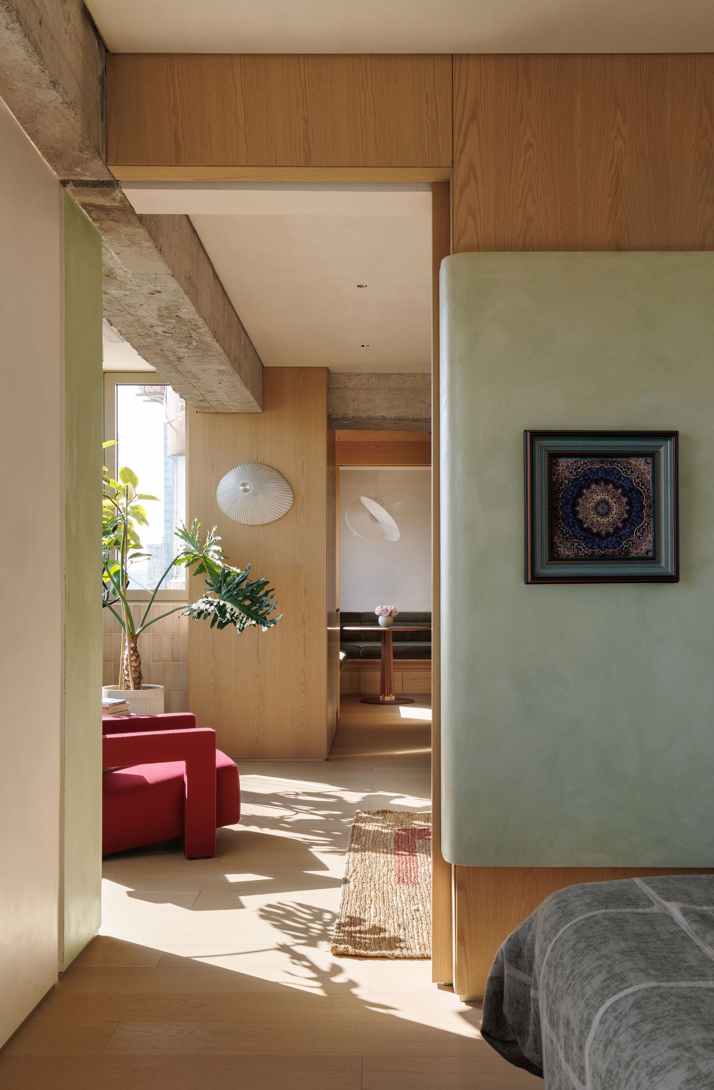 Sunlight streams into a contemporary living space, highlighting the interplay of light and shadow on the pale wooden floors. A green-hued panel with framed art serves as a room divider, complementing the warm wood and exposed concrete beams. A striking red armchair and a lush green plant add vibrant accents to this minimalist and inviting interior. Sunlight streams into a contemporary living space, highlighting the interplay of light and shadow on the pale wooden floors. A green-hued panel with framed art serves as a room divider, complementing the warm wood and exposed concrete beams. A striking red armchair and a lush green plant add vibrant accents to this minimalist and inviting interior.