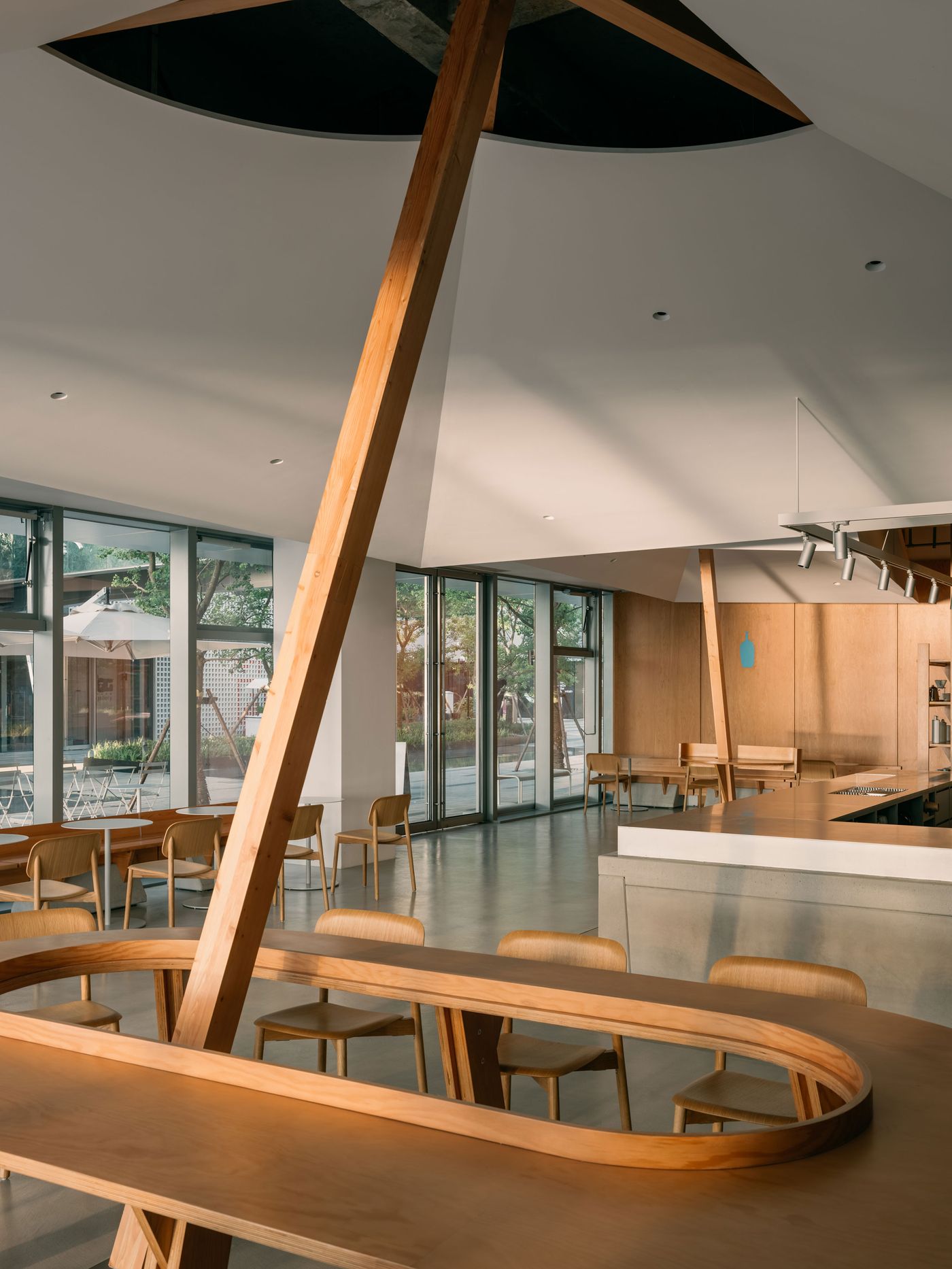 A serene café interior bathed in natural light, featuring asymmetrical ceiling planes, custom timber seating, and a central oval communal table supported by a dramatic leaning wooden beam.
