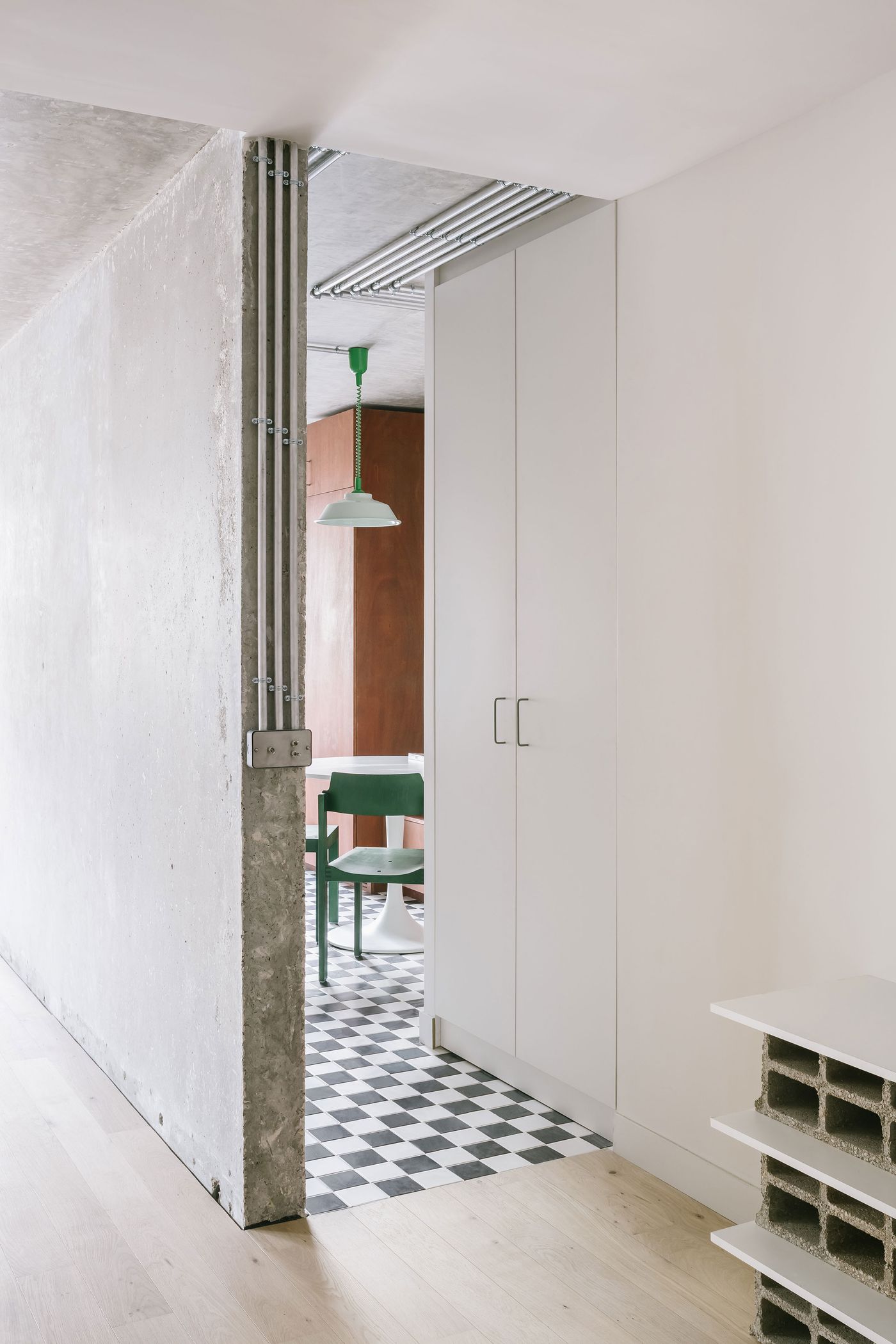 Modern interior space featuring a concrete wall, green pendant light, and a checkered floor leading to a dining area with sleek furniture. Modern interior space featuring a concrete wall, green pendant light, and a checkered floor leading to a dining area with sleek furniture.