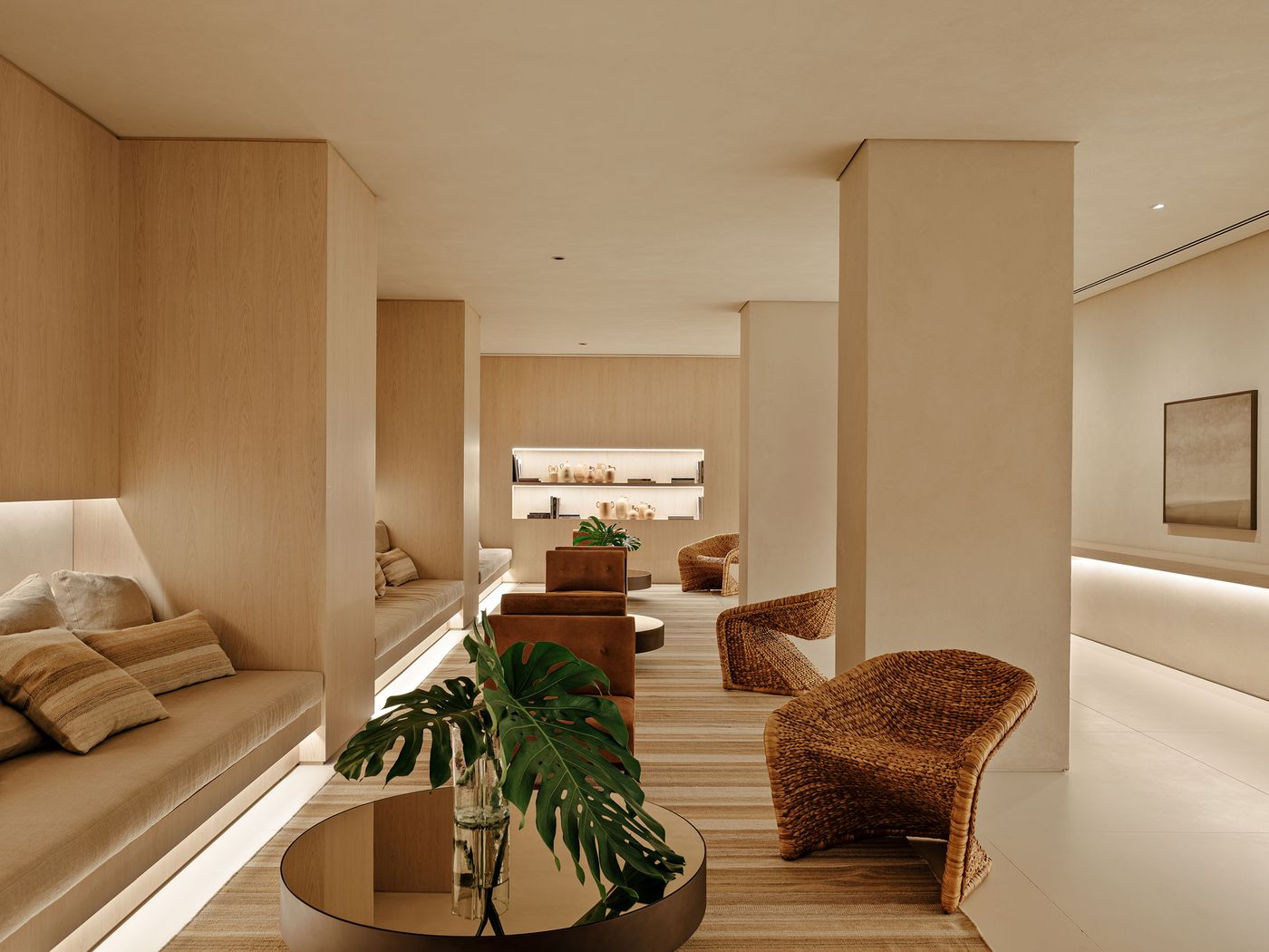 A spacious, minimalist lounge area, bathed in soft, ambient light that highlights the pale wood paneling and natural-toned upholstery. Woven rattan chairs provide organic texture, while a large monstera leaf in a glass vase on a dark circular coffee table introduces a biophilic element. A spacious, minimalist lounge area, bathed in soft, ambient light that highlights the pale wood paneling and natural-toned upholstery. Woven rattan chairs provide organic texture, while a large monstera leaf in a glass vase on a dark circular coffee table introduces a biophilic element.
