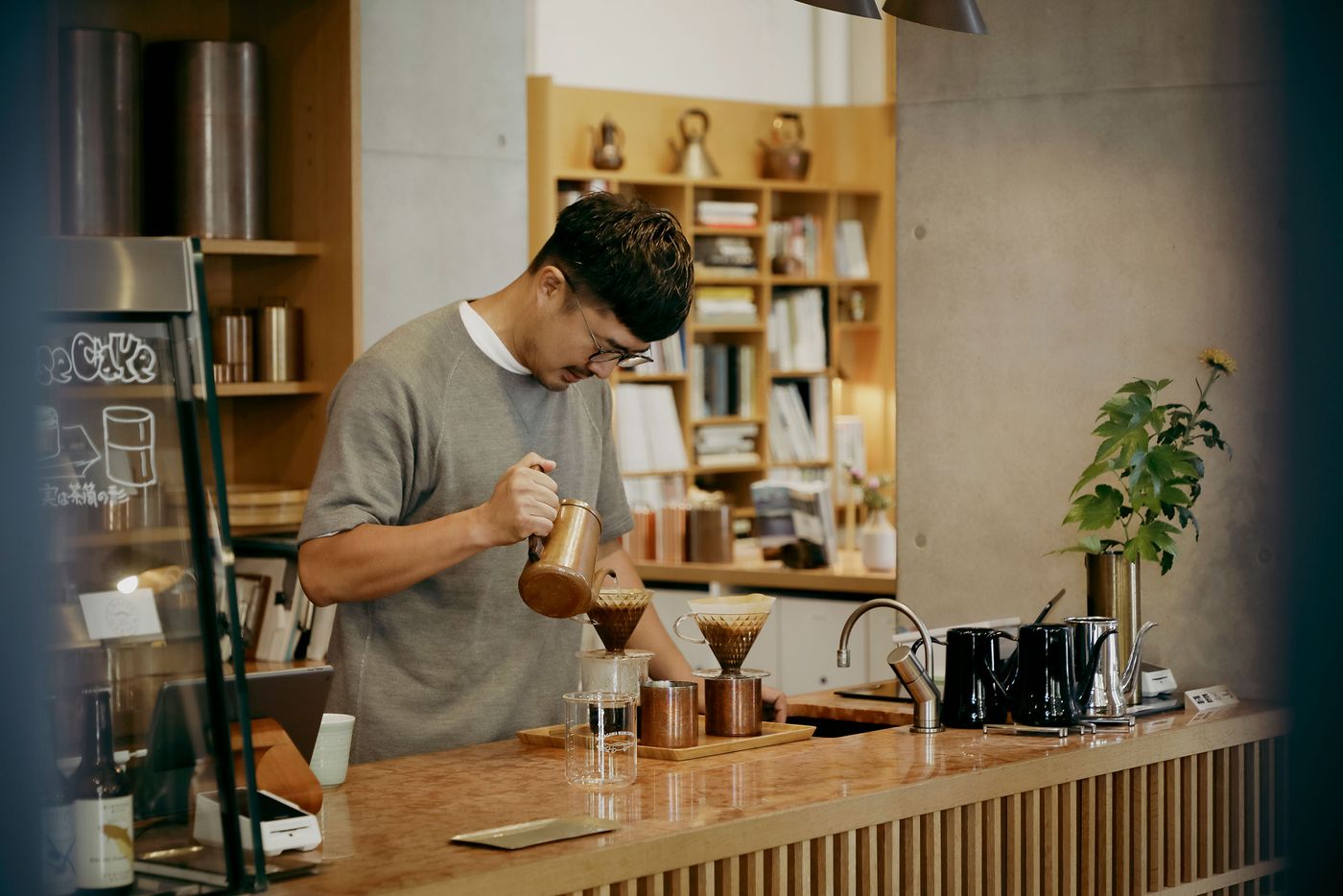 A person pours hot water from a kettle over coffee grounds in a café, surrounded by brewing equipment and a minimalist interior. A person pours hot water from a kettle over coffee grounds in a café, surrounded by brewing equipment and a minimalist interior.