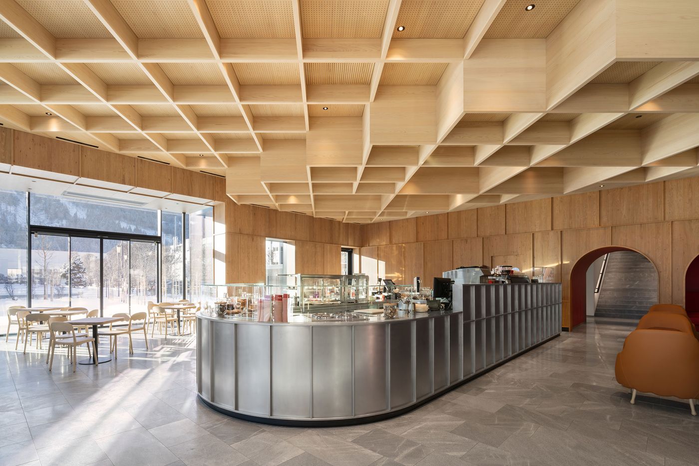 A sophisticated café interior features an undulating coffered wood ceiling with integrated lighting, creating dynamic shadows above a polished concrete floor. The central curved steel counter anchors the space, while floor-to-ceiling windows frame winter mountain vistas, blending Scandinavian minimalism with Alpine sensibility. A sophisticated café interior features an undulating coffered wood ceiling with integrated lighting, creating dynamic shadows above a polished concrete floor. The central curved steel counter anchors the space, while floor-to-ceiling windows frame winter mountain vistas, blending Scandinavian minimalism with Alpine sensibility.