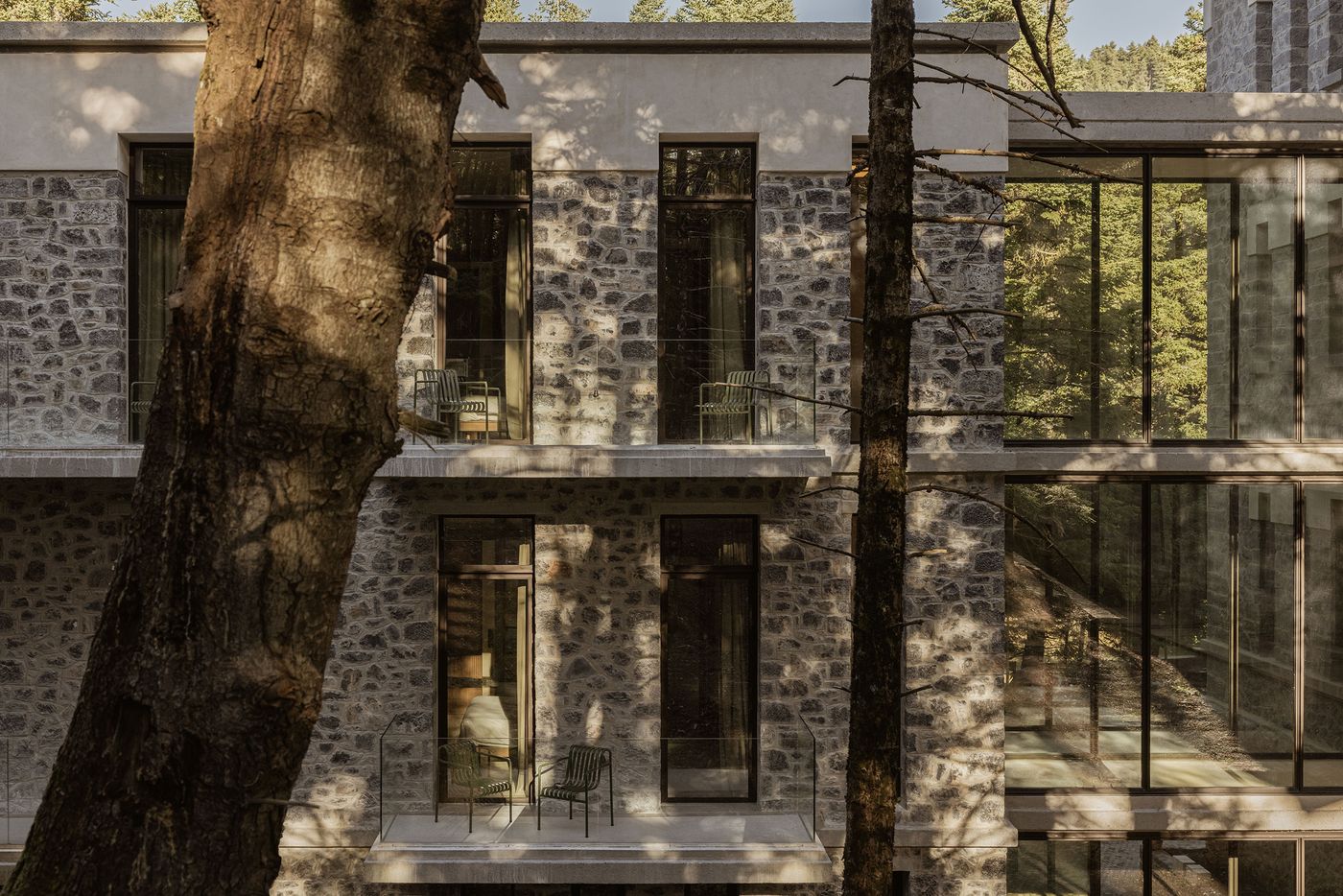 A neo-vernacular hotel facade in the Peloponnese, Greece, showcases robust grey stone masonry contrasted with expansive dark-framed windows. Towering, slender trees in the foreground partially obscure the building, integrating the structure harmoniously with its natural, mountainous surroundings and emphasizing a biophilic design approach. A neo-vernacular hotel facade in the Peloponnese, Greece, showcases robust grey stone masonry contrasted with expansive dark-framed windows. Towering, slender trees in the foreground partially obscure the building, integrating the structure harmoniously with its natural, mountainous surroundings and emphasizing a biophilic design approach.