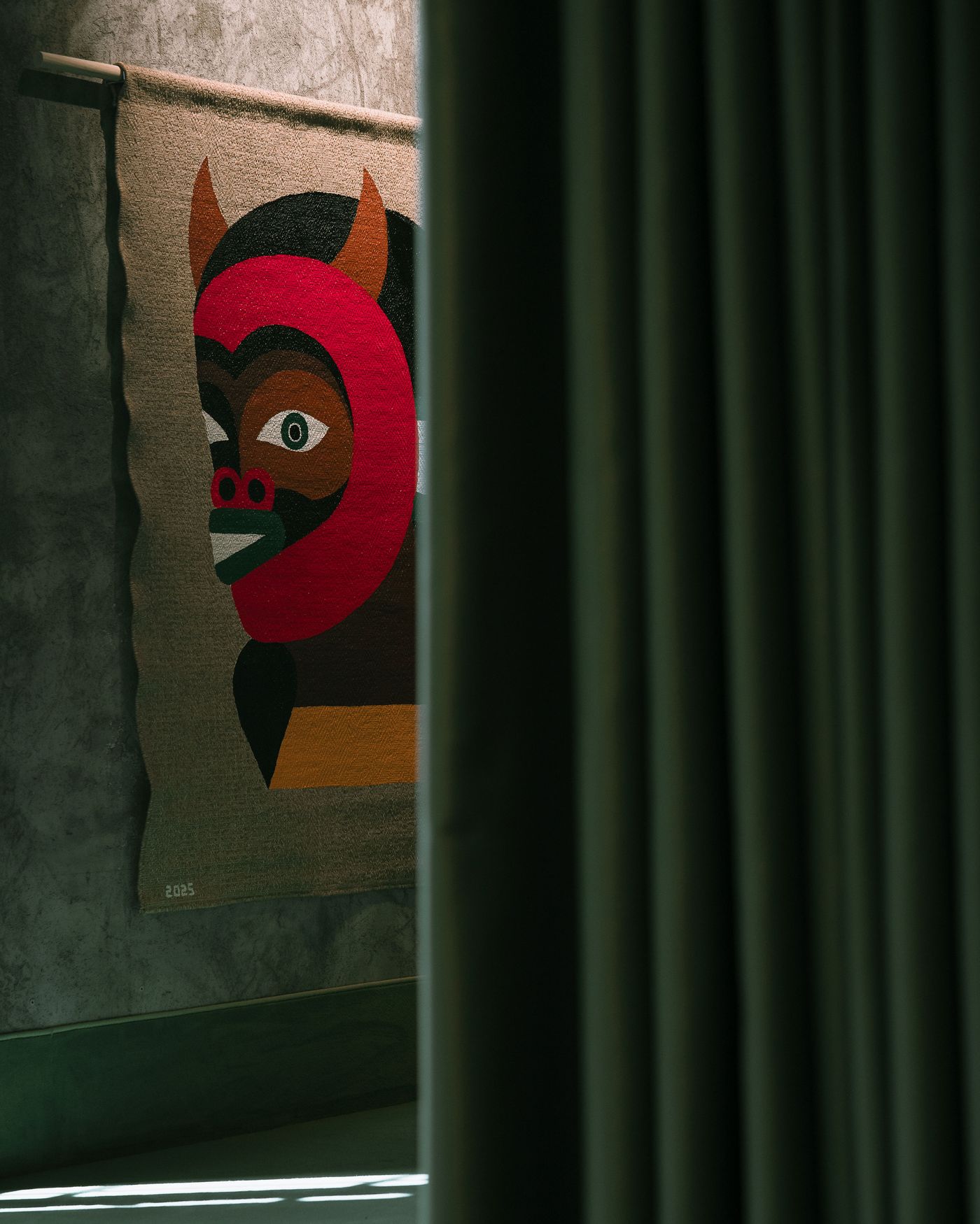 A colorful, abstract textile artwork featuring a stylized figure with horns, partially visible through creased curtains.