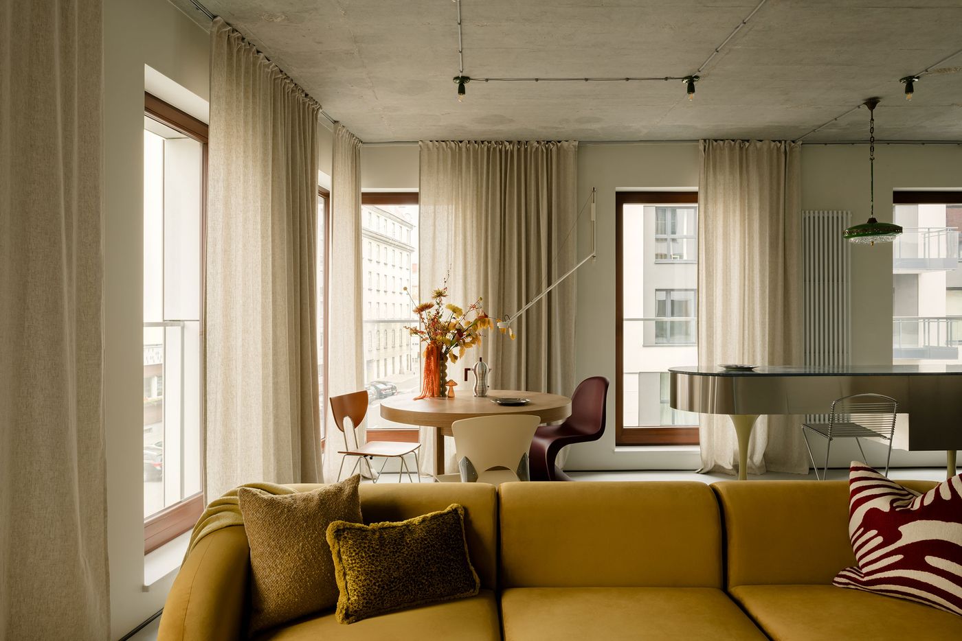 Sunlight floods a corner dining and kitchen area, where industrial concrete ceilings meet full-length curtains and sculptural lighting. A curved mustard sofa and round wooden table introduce warmth, while mismatched dining chairs and a sleek metal island express Mistovia’s eclectic design language. Sunlight floods a corner dining and kitchen area, where industrial concrete ceilings meet full-length curtains and sculptural lighting. A curved mustard sofa and round wooden table introduce warmth, while mismatched dining chairs and a sleek metal island express Mistovia’s eclectic design language.
