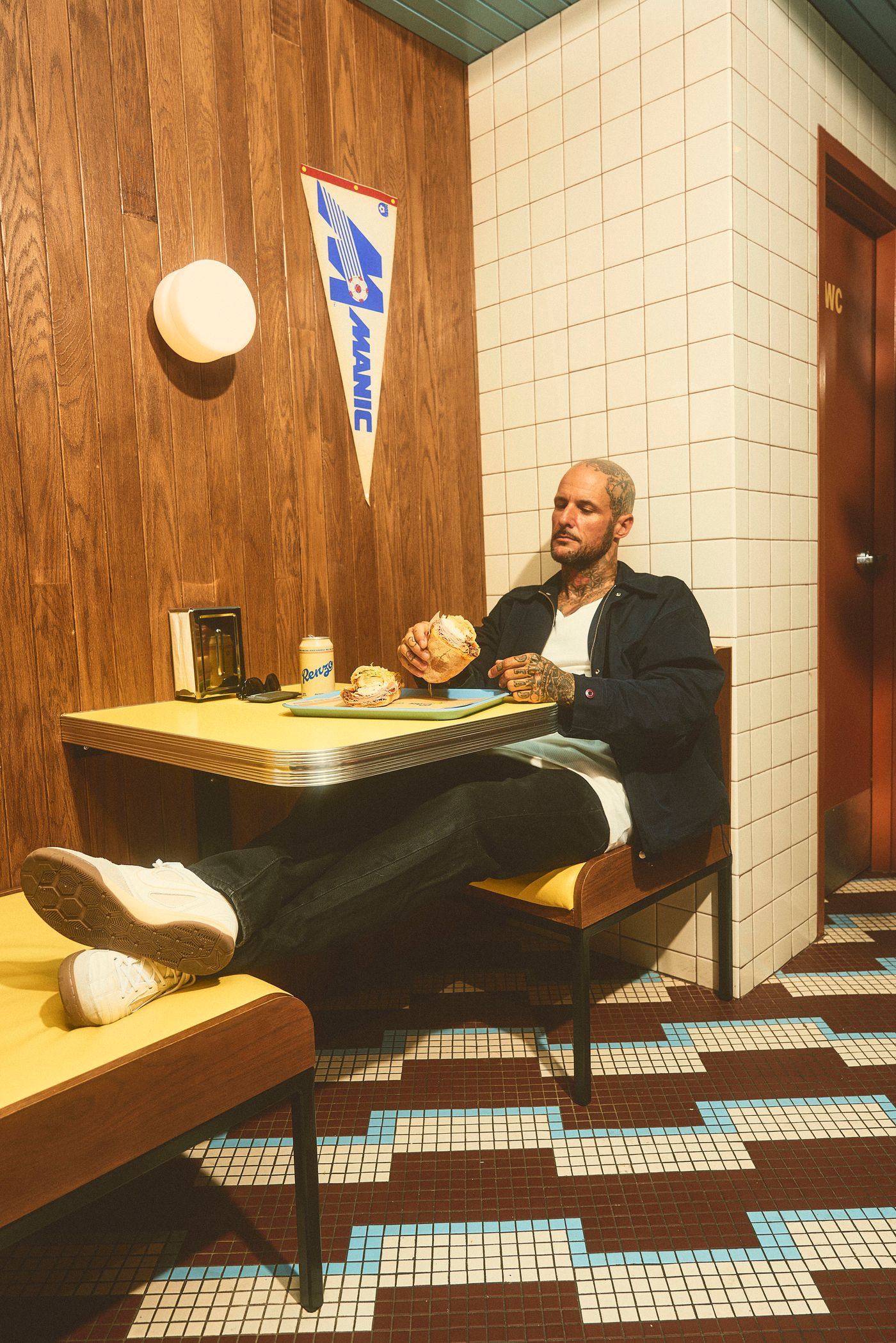 A man with tattoos sits at a yellow-topped diner table, eating a sandwich. The interior features wood-paneled walls, tiled floors in a geometric pattern of brown and blue, and a pennant displaying "MANIC." A small waste bin and a canned beverage sit on the table, evoking a midcentury-modern, retro vibe.