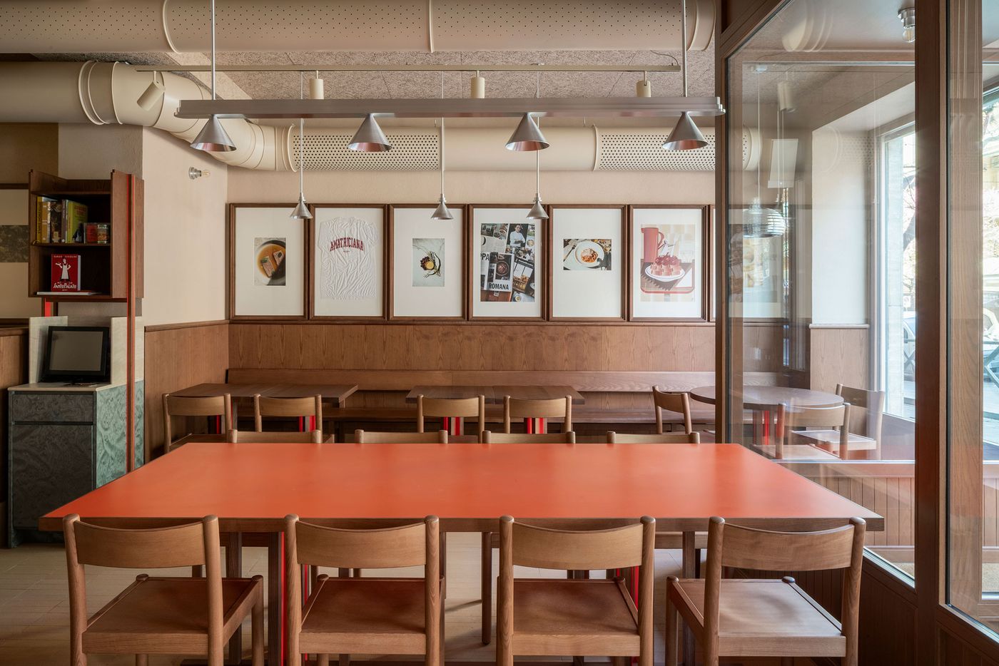 An inviting trattoria interior by Naessi Studio for Santo Palato in Rome. A vibrant orange communal table, accented with red legs, anchors the foreground. Behind it, a row of framed art hangs above banquette seating and simple wooden tables, set against warm wood paneling and exposed industrial-style ceiling pipes with minimalist pendant lights. An inviting trattoria interior by Naessi Studio for Santo Palato in Rome. A vibrant orange communal table, accented with red legs, anchors the foreground. Behind it, a row of framed art hangs above banquette seating and simple wooden tables, set against warm wood paneling and exposed industrial-style ceiling pipes with minimalist pendant lights.