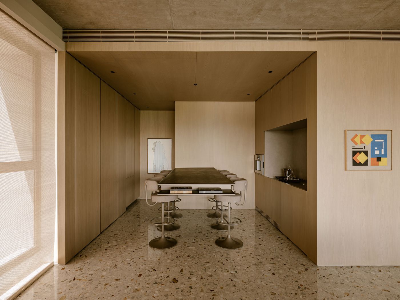 Modern interior featuring a minimalist kitchen and dining area with a long table, sleek stools, wooden panels, and colorful artwork. Modern interior featuring a minimalist kitchen and dining area with a long table, sleek stools, wooden panels, and colorful artwork.