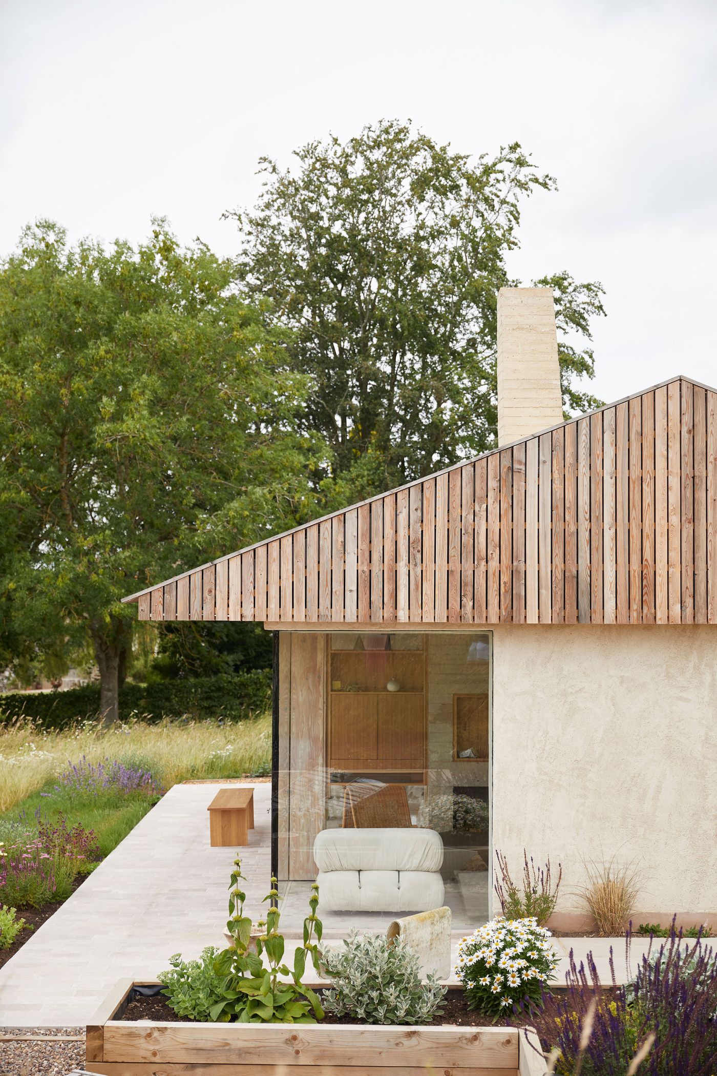 The Maker's Barn by Hutch Design showcases a striking blend of neo-vernacular and minimalist styles. A pitched roof with vertical timber cladding meets a smooth, pale facade and an expansive corner of glass, revealing a glimpse of minimalist interior furnishings. Thoughtfully integrated garden beds with lush greenery and white flowers connect the architecture to its natural surroundings. The Maker's Barn by Hutch Design showcases a striking blend of neo-vernacular and minimalist styles. A pitched roof with vertical timber cladding meets a smooth, pale facade and an expansive corner of glass, revealing a glimpse of minimalist interior furnishings. Thoughtfully integrated garden beds with lush greenery and white flowers connect the architecture to its natural surroundings.