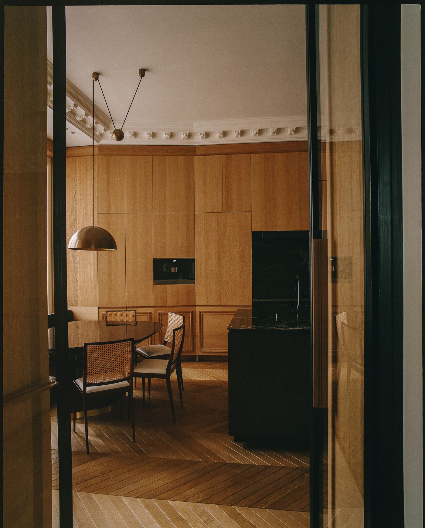 A warm, inviting kitchen and dining area with exquisite oak paneling enveloping the walls, creating a cohesive, built-in aesthetic. A minimalist black island anchors the space, while a circular dining table with cane-back chairs and a brass pendant light introduces a touch of vintage-retro elegance, all set on a herringbone wood floor. A warm, inviting kitchen and dining area with exquisite oak paneling enveloping the walls, creating a cohesive, built-in aesthetic. A minimalist black island anchors the space, while a circular dining table with cane-back chairs and a brass pendant light introduces a touch of vintage-retro elegance, all set on a herringbone wood floor.