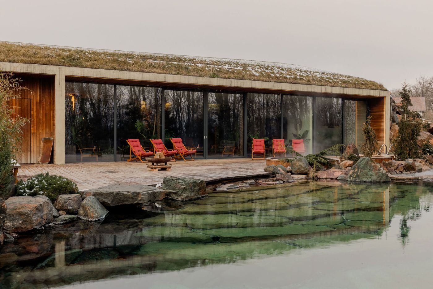Modern house with a grassy roof, large glass walls, and wooden deck, featuring red loungers beside a tranquil, rocky pond. Modern house with a grassy roof, large glass walls, and wooden deck, featuring red loungers beside a tranquil, rocky pond.