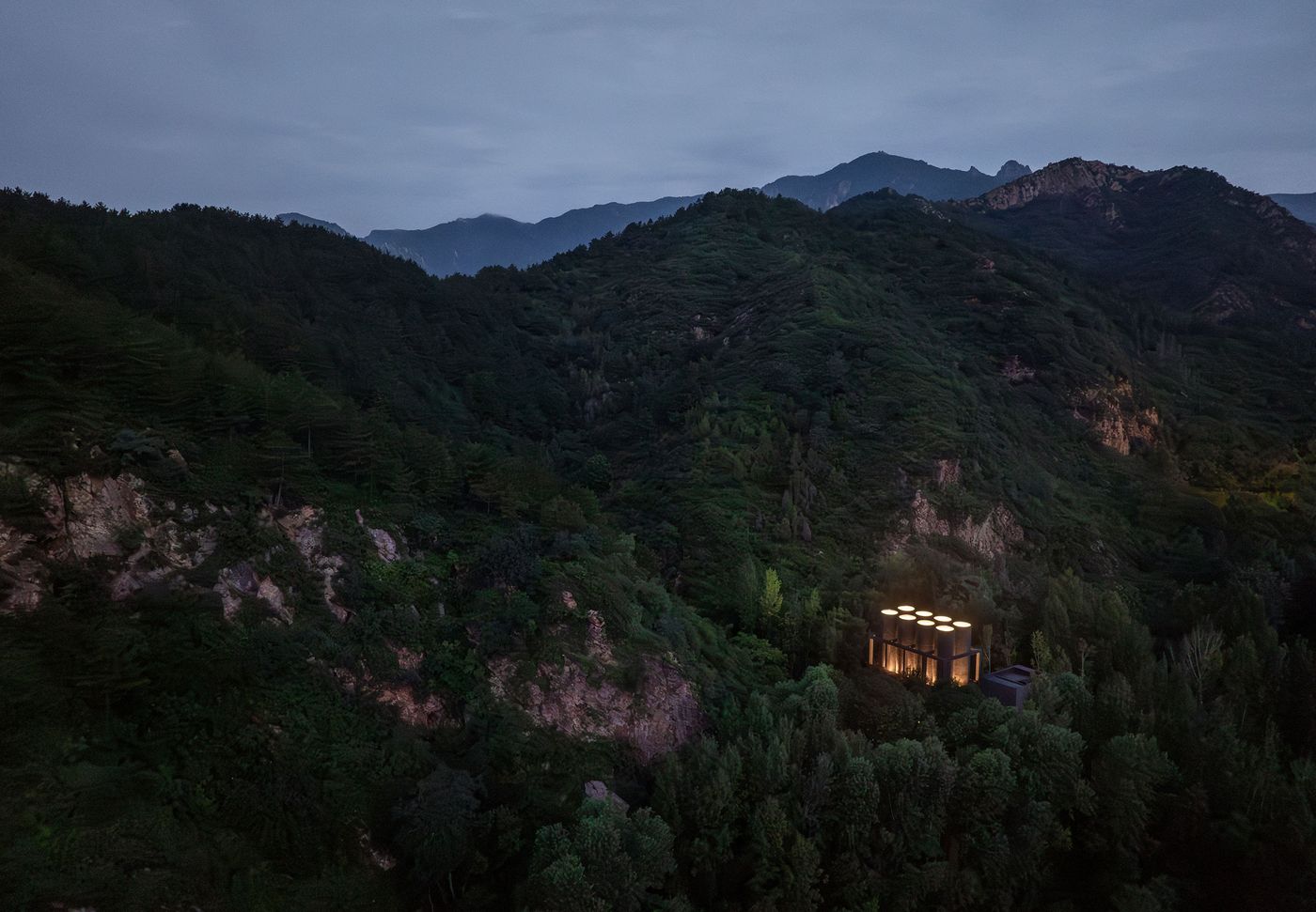 A modern building with illuminated circular roofs stands amidst lush, dark green mountains under a dusk sky. A modern building with illuminated circular roofs stands amidst lush, dark green mountains under a dusk sky.