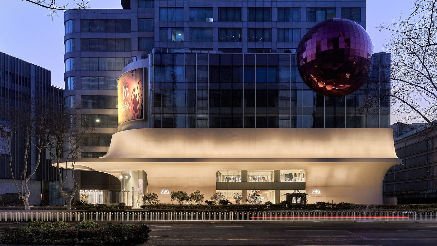 Modern Zara store with a distinctive curved facade and a large, reflective red sphere on top, illuminated at dusk. Modern Zara store with a distinctive curved facade and a large, reflective red sphere on top, illuminated at dusk.