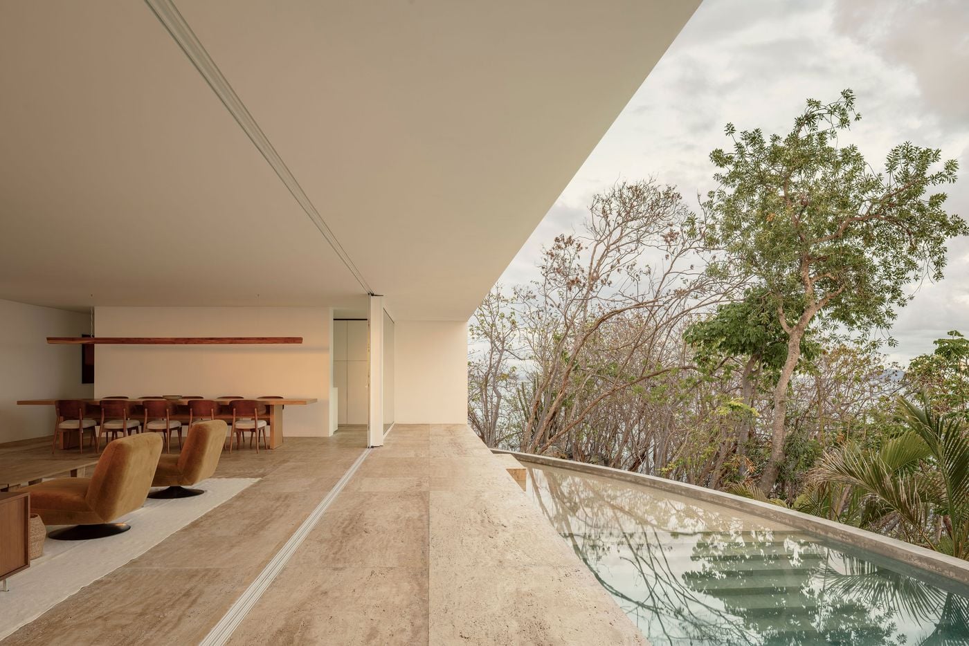 Viewed along the open living level, a long timber dining table, matching chairs with cream cushions and a linear wooden pendant recede into the white interior. To the right, a narrow pool traces a gentle concrete curve along the terrace edge, its surface reflecting the dry forest beyond the cantilevered roof. Viewed along the open living level, a long timber dining table, matching chairs with cream cushions and a linear wooden pendant recede into the white interior. To the right, a narrow pool traces a gentle concrete curve along the terrace edge, its surface reflecting the dry forest beyond the cantilevered roof.
