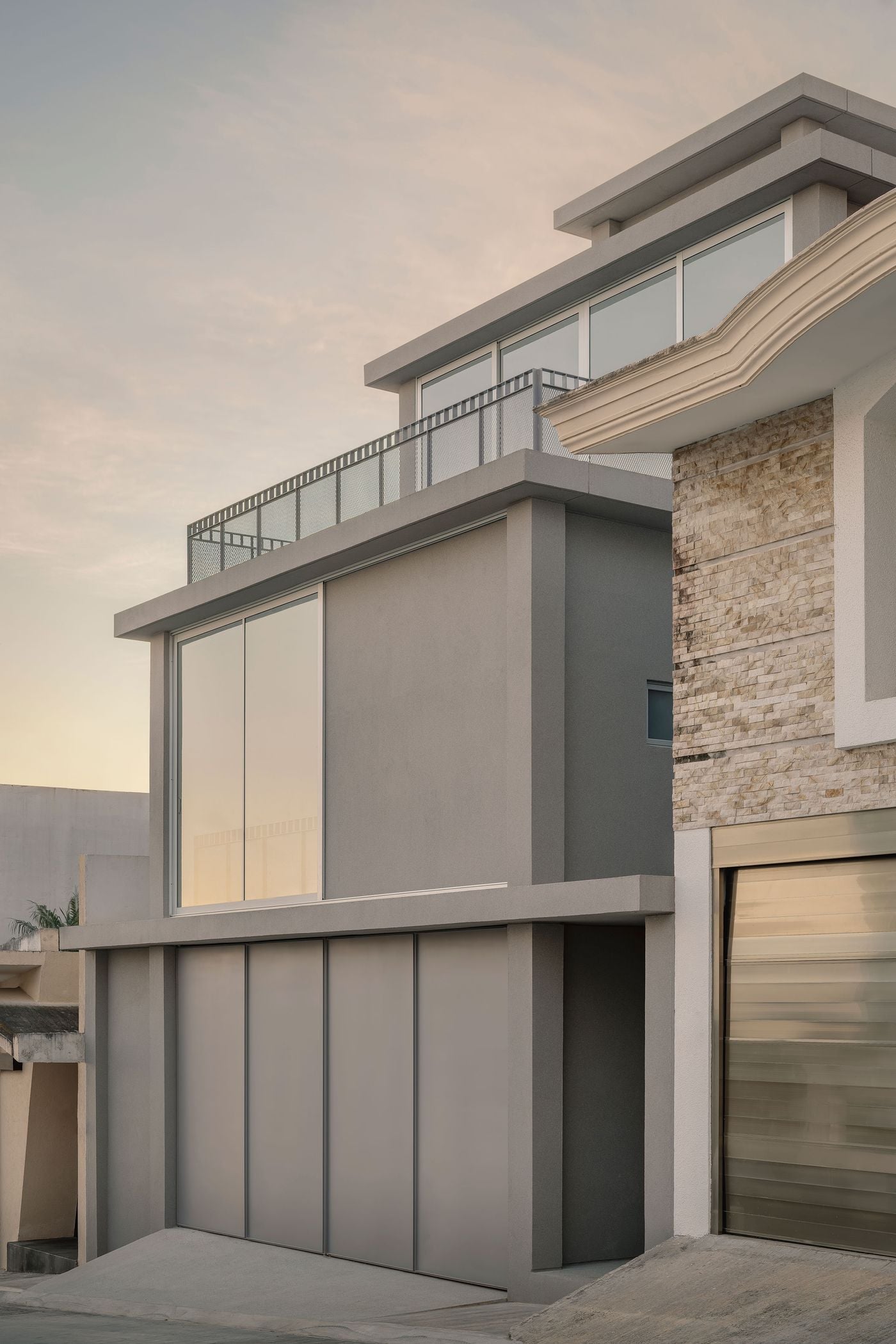 A close street view emphasises Casa Tlaloc’s stepped composition, with grey cubic volumes receding upwards behind projecting slabs and fine metal-mesh balustrades. Expansive glazing reflects the soft evening light, while the house’s restrained geometry forms a marked contrast with the more ornate stone-clad residence next door. A close street view emphasises Casa Tlaloc’s stepped composition, with grey cubic volumes receding upwards behind projecting slabs and fine metal-mesh balustrades. Expansive glazing reflects the soft evening light, while the house’s restrained geometry forms a marked contrast with the more ornate stone-clad residence next door.