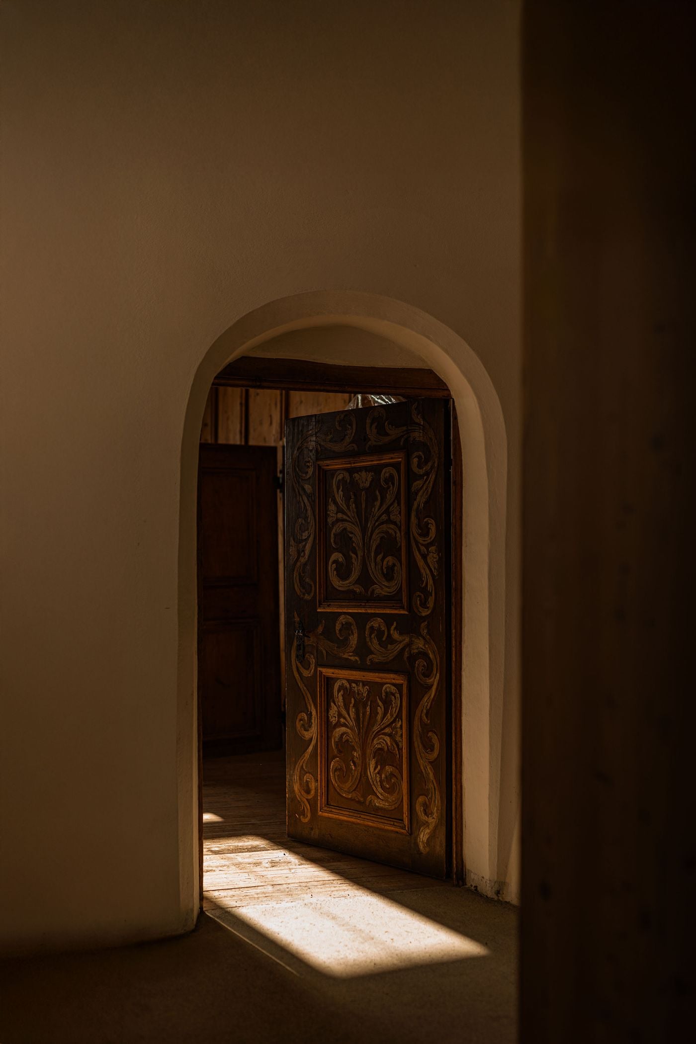 A softly rounded, hand-plastered archway at Castel Badia opens onto a hand-painted timber door decorated with scrolling foliate motifs. A shaft of sunlight falls across worn floorboards into a timber-panelled room beyond. The scene is a quiet study in texture, shadow and traditional Tyrolean craftsmanship. A softly rounded, hand-plastered archway at Castel Badia opens onto a hand-painted timber door decorated with scrolling foliate motifs. A shaft of sunlight falls across worn floorboards into a timber-panelled room beyond. The scene is a quiet study in texture, shadow and traditional Tyrolean craftsmanship.