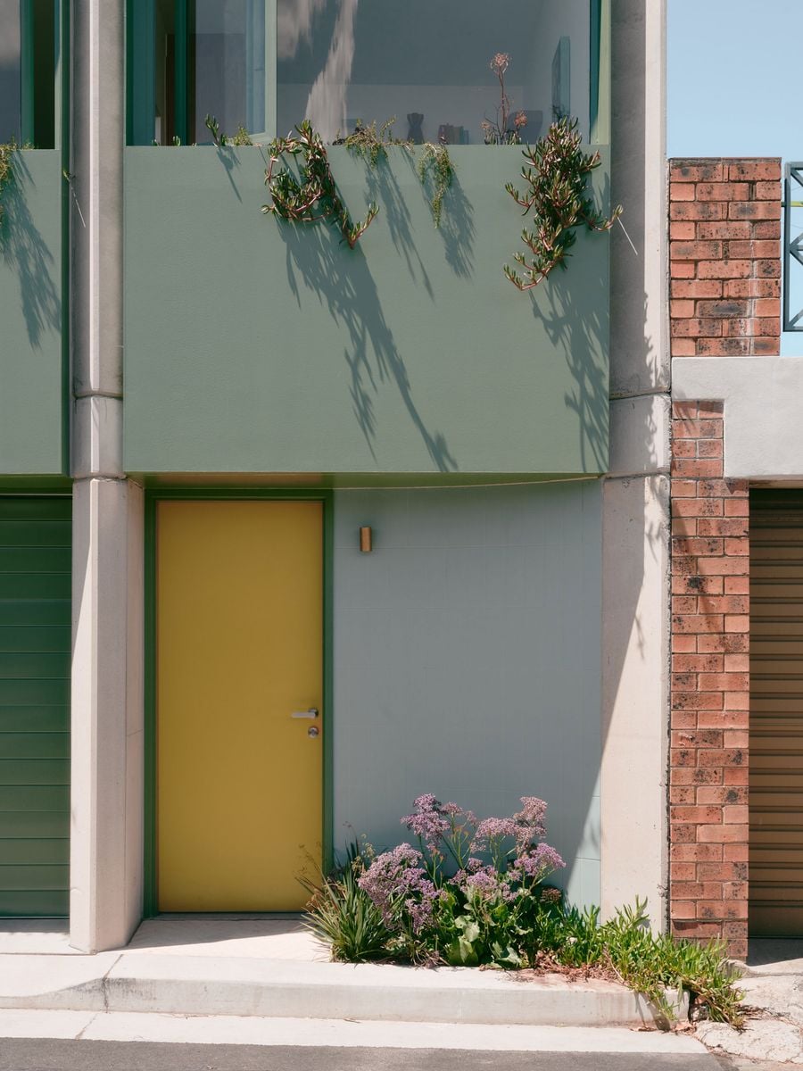 A close view of the entrance reveals the project’s layered palette of sage green, mustard yellow, pale concrete and red brick. Trailing succulents spill over the upper planter, casting crisp shadows in the sun, while a small garden bed at pavement level softens the threshold between architecture and laneway. A close view of the entrance reveals the project’s layered palette of sage green, mustard yellow, pale concrete and red brick. Trailing succulents spill over the upper planter, casting crisp shadows in the sun, while a small garden bed at pavement level softens the threshold between architecture and laneway.