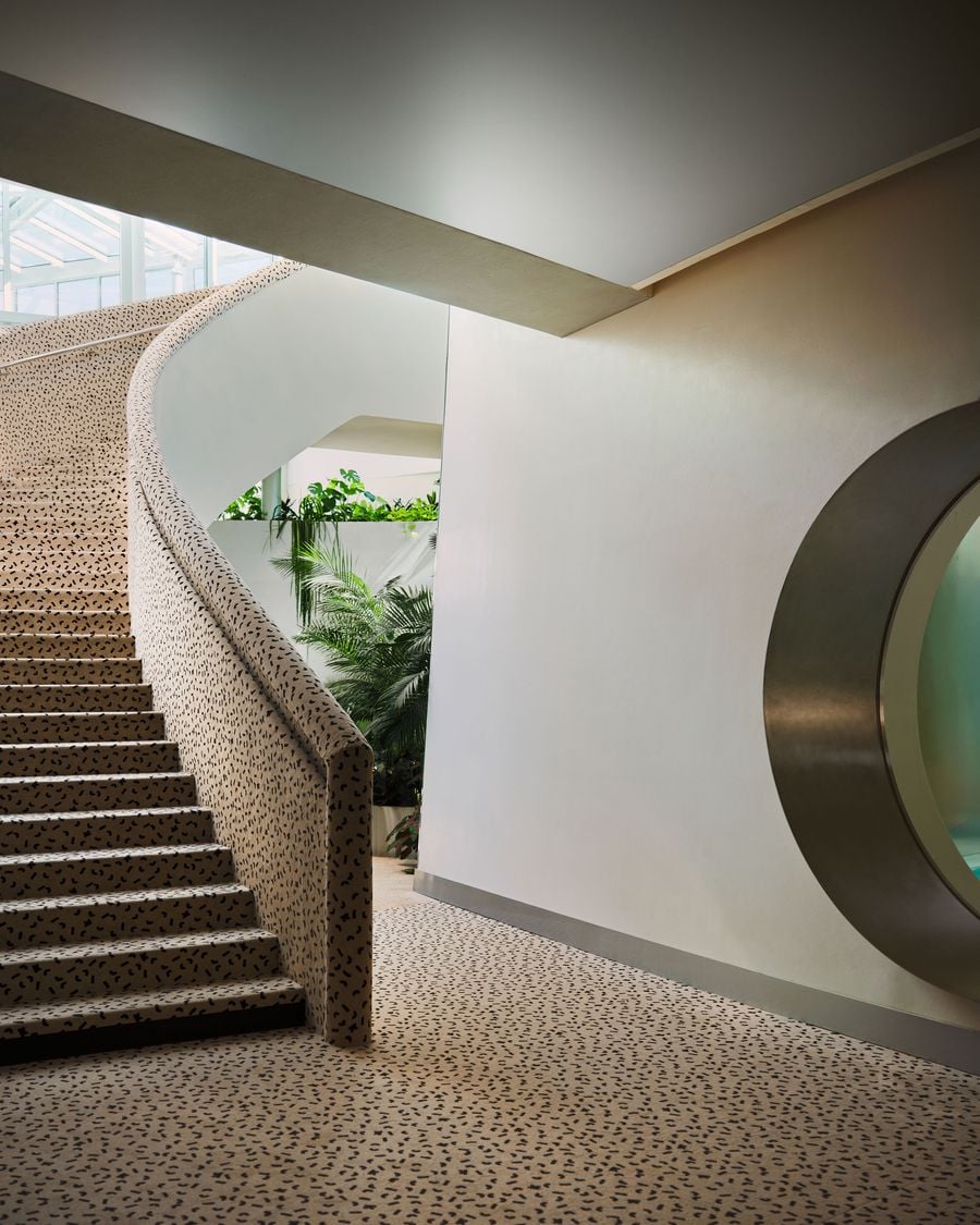 The Ken Club’s main staircase curves upward beside a large circular opening edged in metal, one of several porthole-like motifs threaded through the interior. Cream-and-black flecked upholstery continues from the stair onto the floor, while planting glimpsed beyond softens the otherwise sculptural composition. The Ken Club’s main staircase curves upward beside a large circular opening edged in metal, one of several porthole-like motifs threaded through the interior. Cream-and-black flecked upholstery continues from the stair onto the floor, while planting glimpsed beyond softens the otherwise sculptural composition.