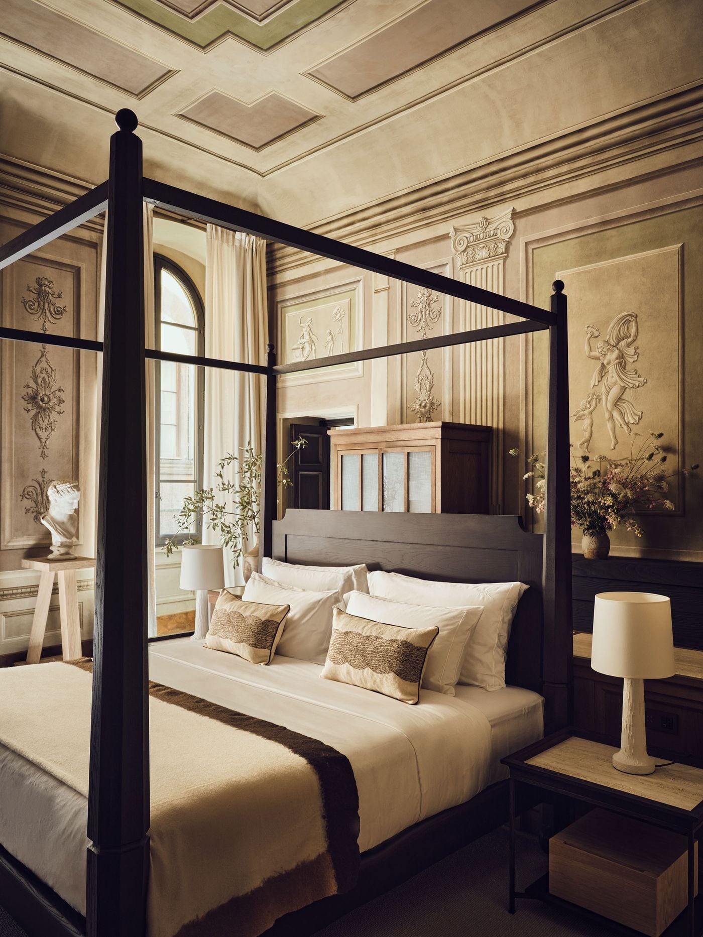 A bedroom at La Réserve Firenze enveloped in restored painted decoration: trompe-l'oeil pilasters, ornamental panels and dancing classical figures in muted stone tones. A dark-stained four-poster bed, a classical bust and a linen-panelled cabinet introduce a contemporary restraint that lets the room's painted grandeur lead. A bedroom at La Réserve Firenze enveloped in restored painted decoration: trompe-l'oeil pilasters, ornamental panels and dancing classical figures in muted stone tones. A dark-stained four-poster bed, a classical bust and a linen-panelled cabinet introduce a contemporary restraint that lets the room's painted grandeur lead.