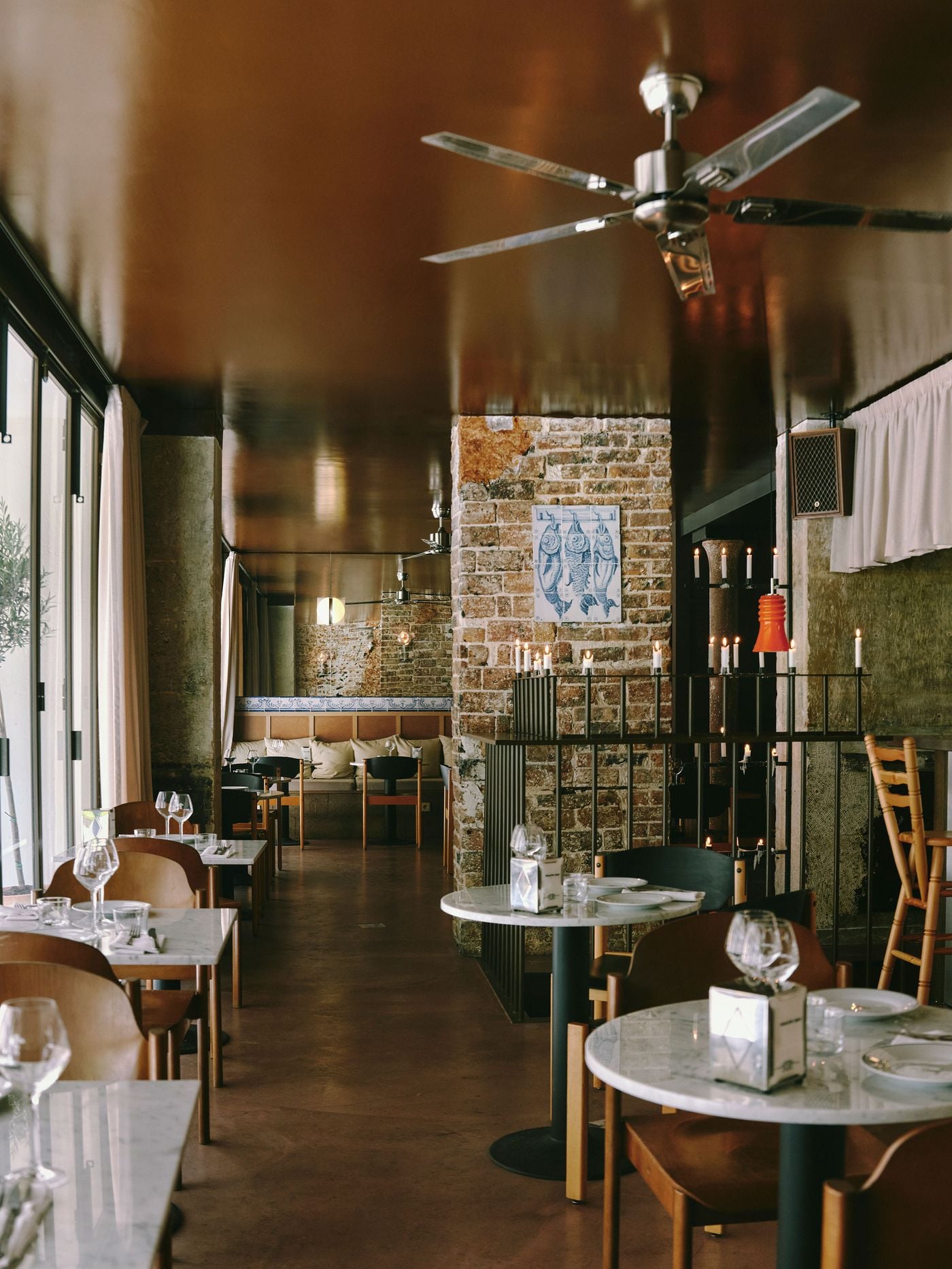 A vertical perspective of the SaltoBar dining room in Paris, crowned by a steel ceiling fan against a glossy copper-brown ceiling. Marble-topped tables lead the eye past raw concrete and brick piers to a banquette trimmed with a blue-and-white tile frieze. A fish-motif azulejo panel and candlelit railing punctuate the right. A vertical perspective of the SaltoBar dining room in Paris, crowned by a steel ceiling fan against a glossy copper-brown ceiling. Marble-topped tables lead the eye past raw concrete and brick piers to a banquette trimmed with a blue-and-white tile frieze. A fish-motif azulejo panel and candlelit railing punctuate the right.
