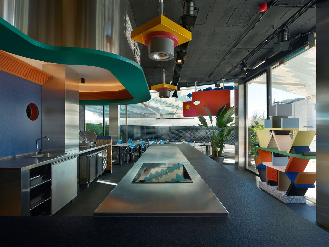 A long stainless-steel kitchen island at Seth & Manni's leads the eye towards glazed walls and a zigzag-patterned awning. A teal overhang curves above, and chunky yellow and red pendant lights hang from the black ceiling. Ettore Sottsass's Carlton bookcase stands by the window like a totem of Memphis geometry. A long stainless-steel kitchen island at Seth & Manni's leads the eye towards glazed walls and a zigzag-patterned awning. A teal overhang curves above, and chunky yellow and red pendant lights hang from the black ceiling. Ettore Sottsass's Carlton bookcase stands by the window like a totem of Memphis geometry.