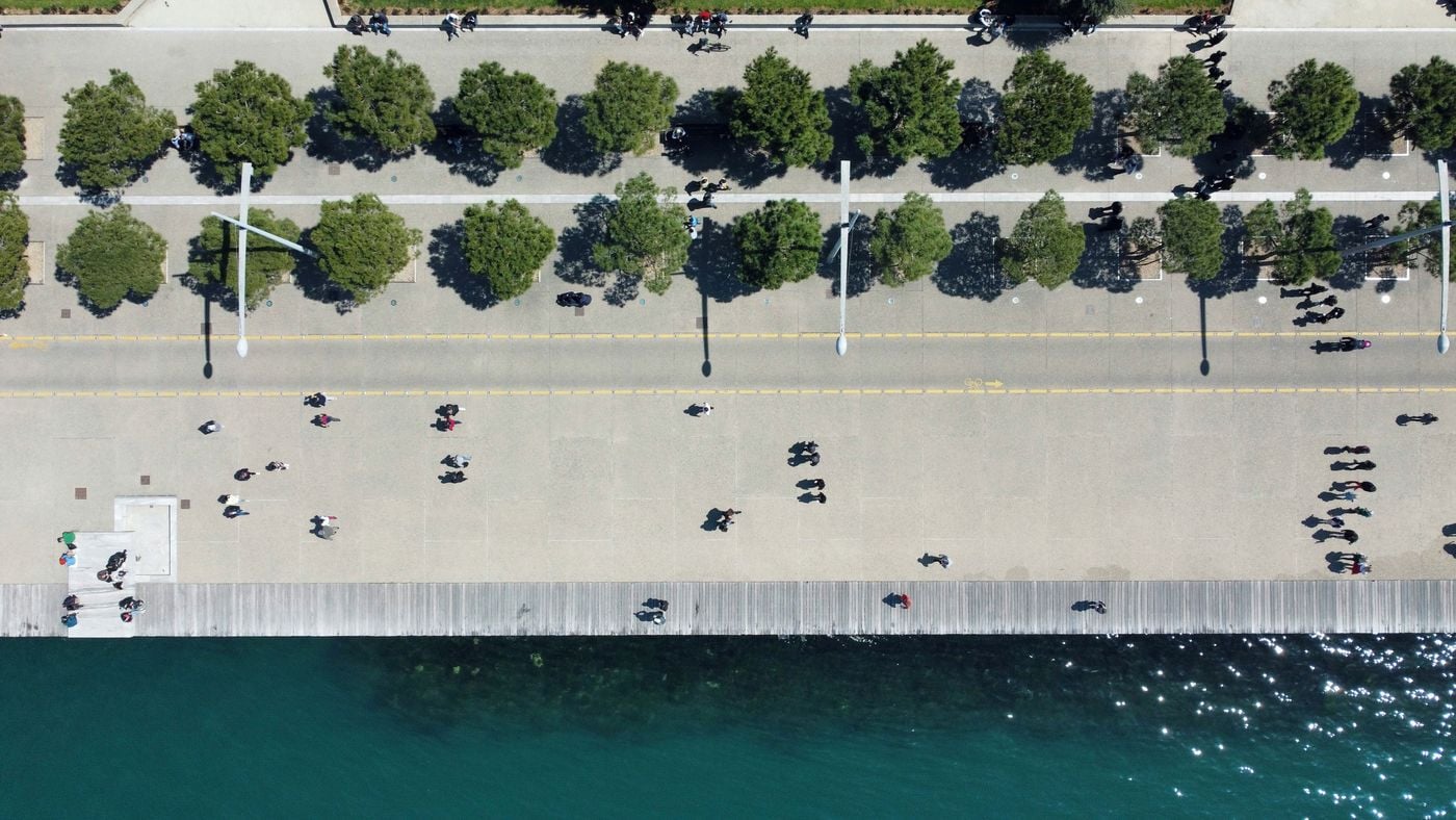 Aerial view of Thessaloniki’s New Waterfront (Nea Paralia): rows of pine trees, a cycle lane and a timber boardwalk run parallel to the turquoise Thermaic Gulf, pedestrians casting long shadows in strong Mediterranean sunlight.