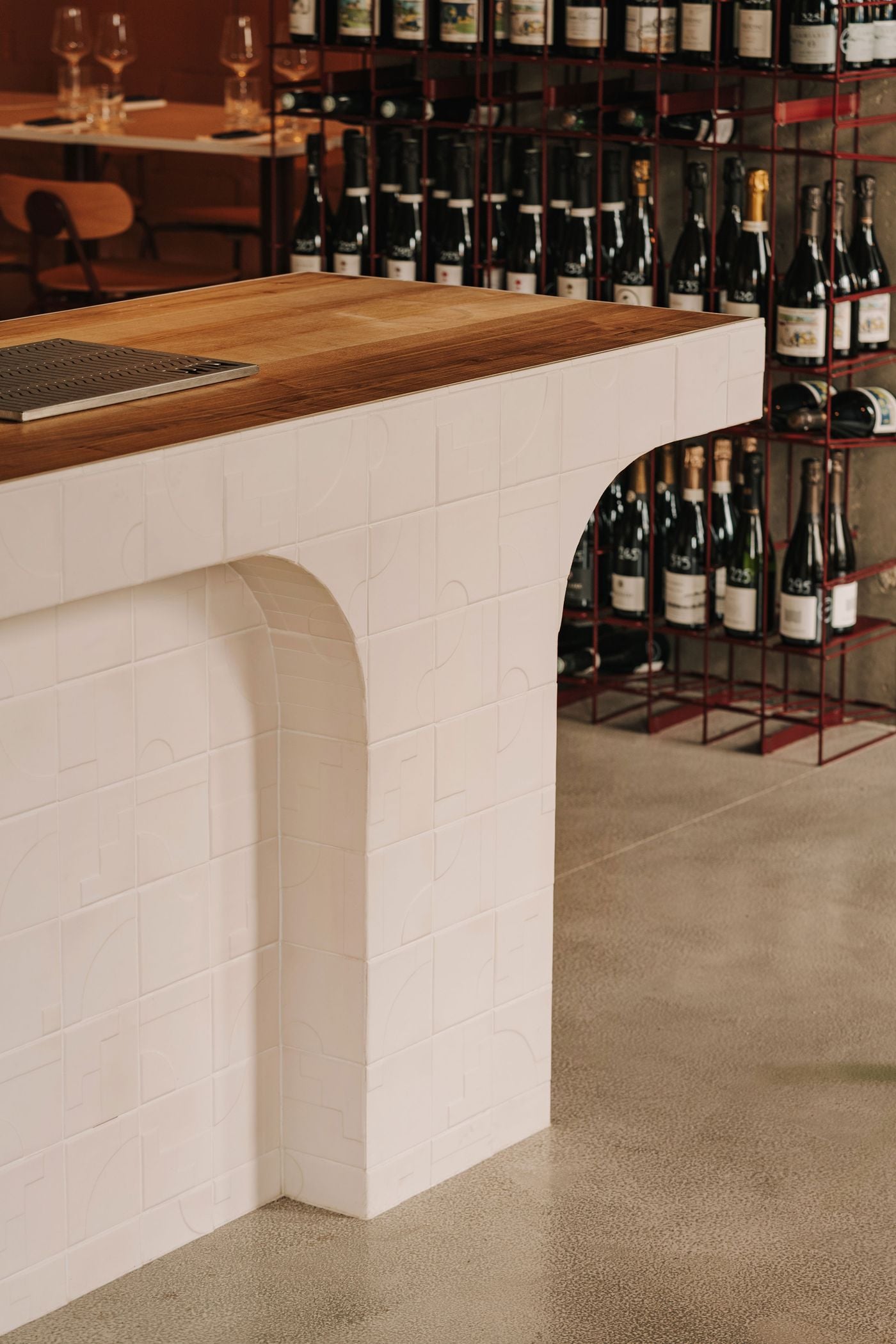 A close-up of WIN's champagne bar counter highlights the contrast between its warm timber top and white glazed ceramic tiles embossed with subtle geometric motifs. A softly curved recess interrupts the counter's monolithic form, introducing an organic element that echoes the sinuous metalwork found throughout the Warsaw interior. A close-up of WIN's champagne bar counter highlights the contrast between its warm timber top and white glazed ceramic tiles embossed with subtle geometric motifs. A softly curved recess interrupts the counter's monolithic form, introducing an organic element that echoes the sinuous metalwork found throughout the Warsaw interior.